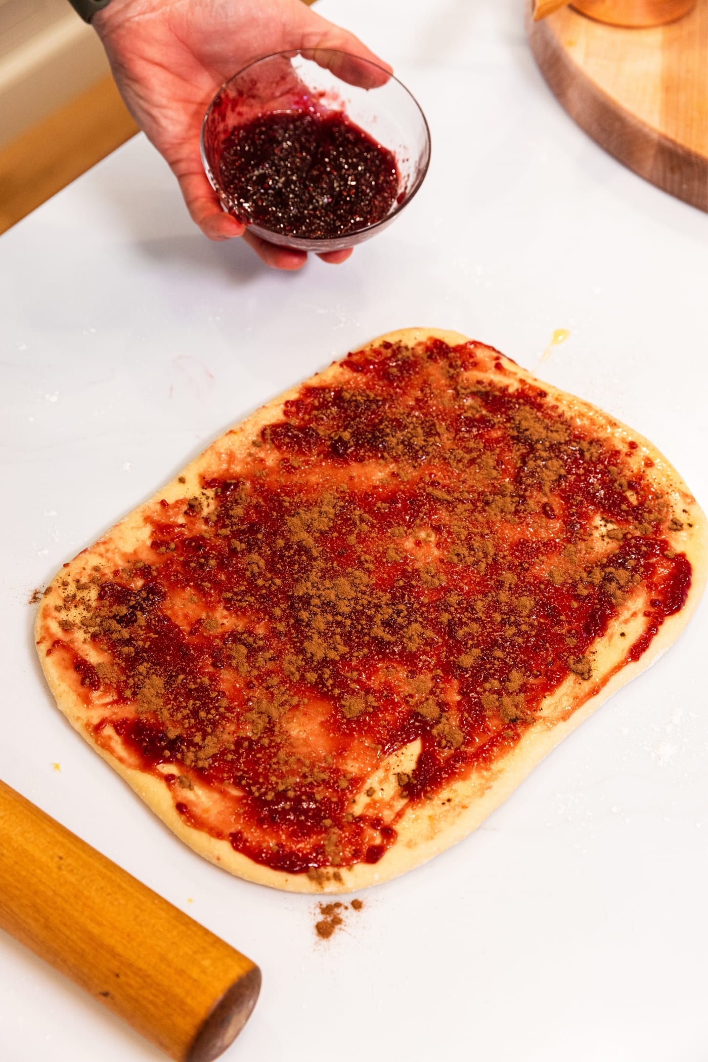 Hand holding small glass bowl filled with raspberry jam after smoothing jam over the surface of unbaked dough rolled into a rectangle with cinnamon sprinkled all over the top as well.