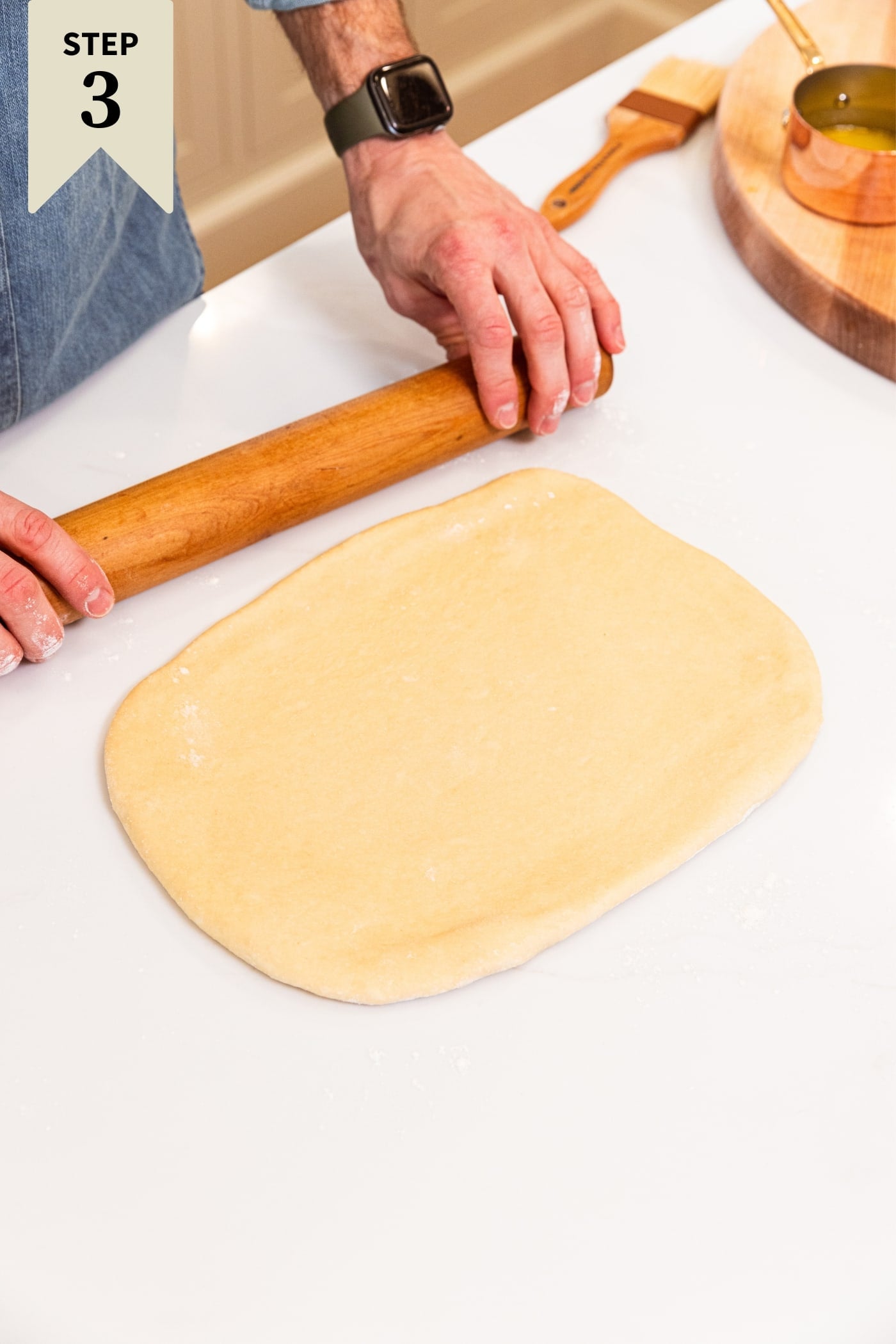 Hand using wooden rolling pin to roll out dough into a large rectangle on white surface with wood cutting board in background.