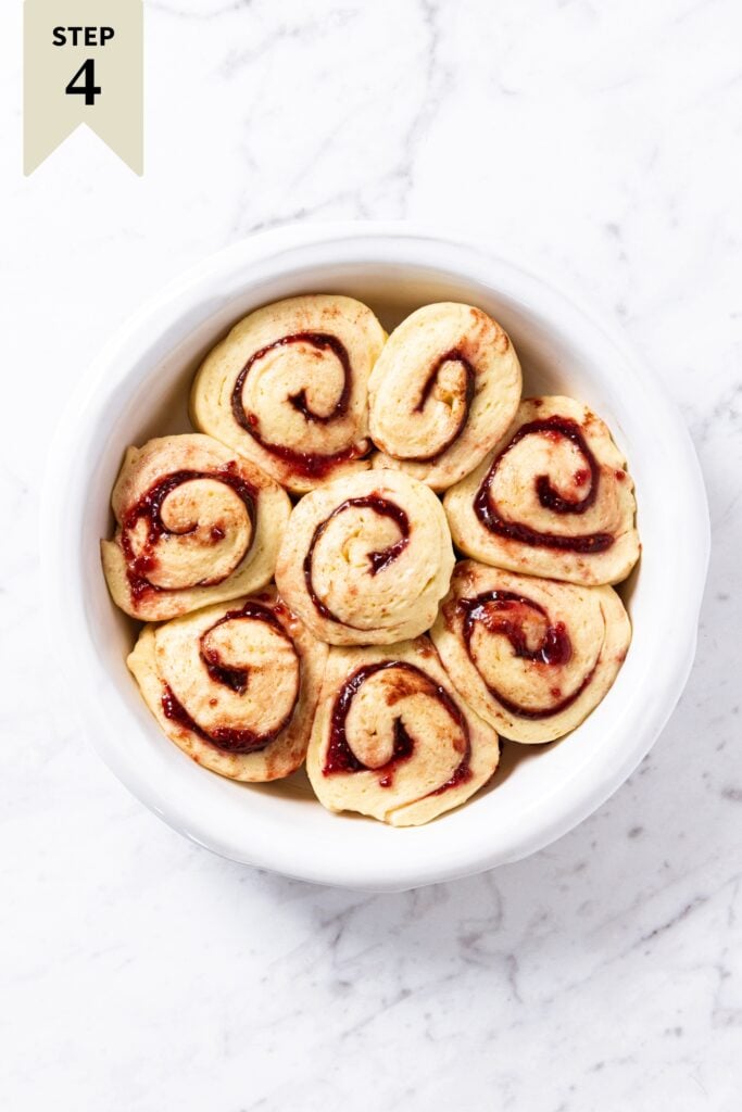 Round baking dish sitting on white marble surface with risen raspberry cinnamon rolls inside right before going into the oven to bake.