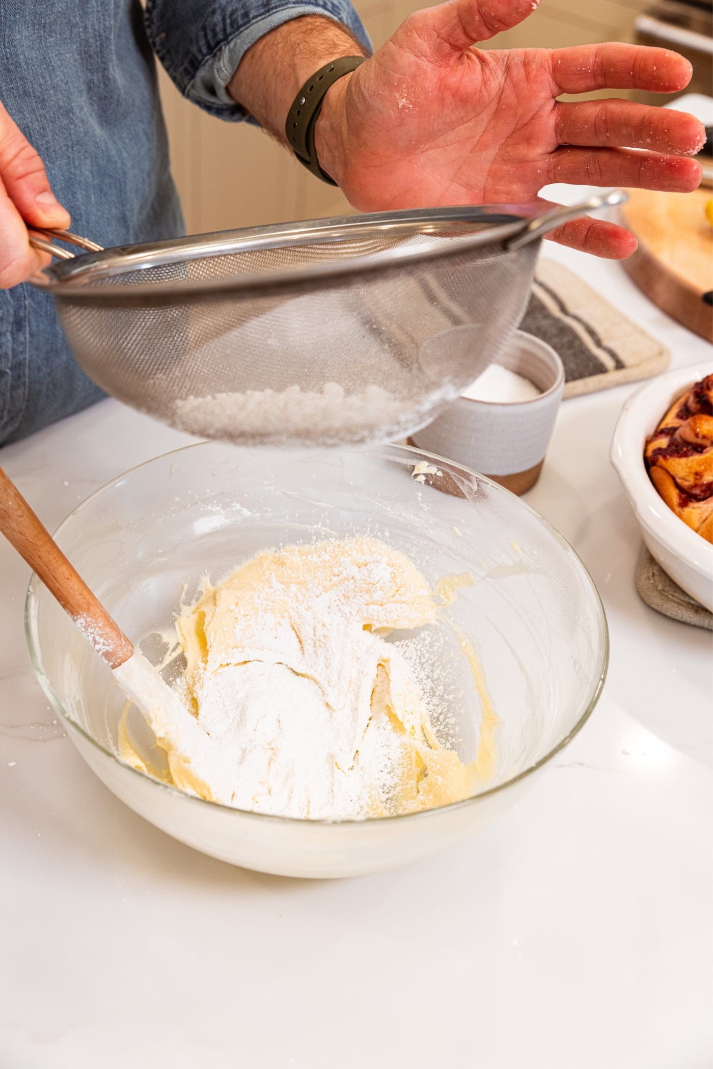 Hand holding strainer using it to sift powdered sugar into a glass bowl already filled with smooth cream cheese on white surface.