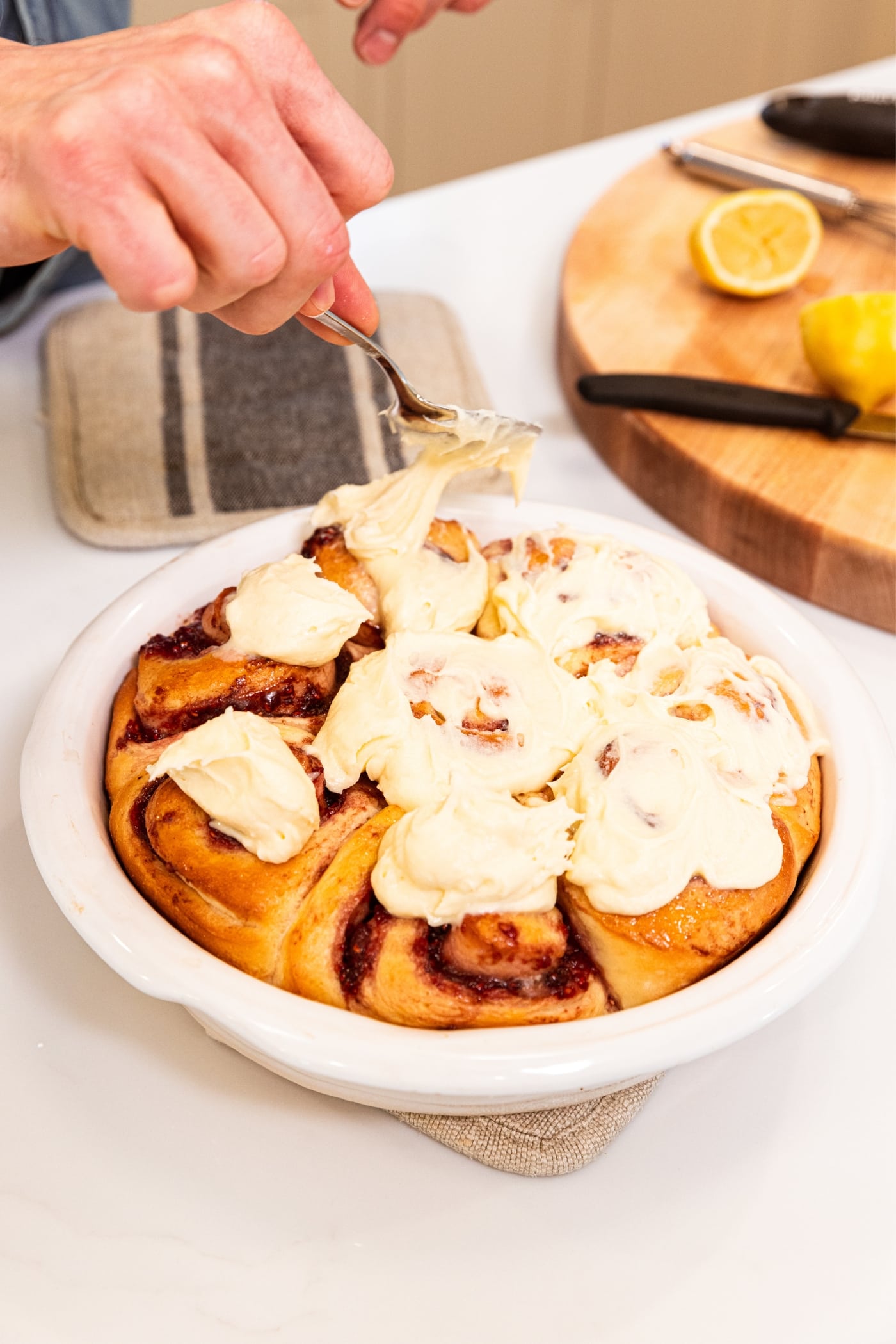 Hand holding spoon with cream cheese frosting dotting the top of raspberry cinnamon rolls before spreading it out to cover surface.