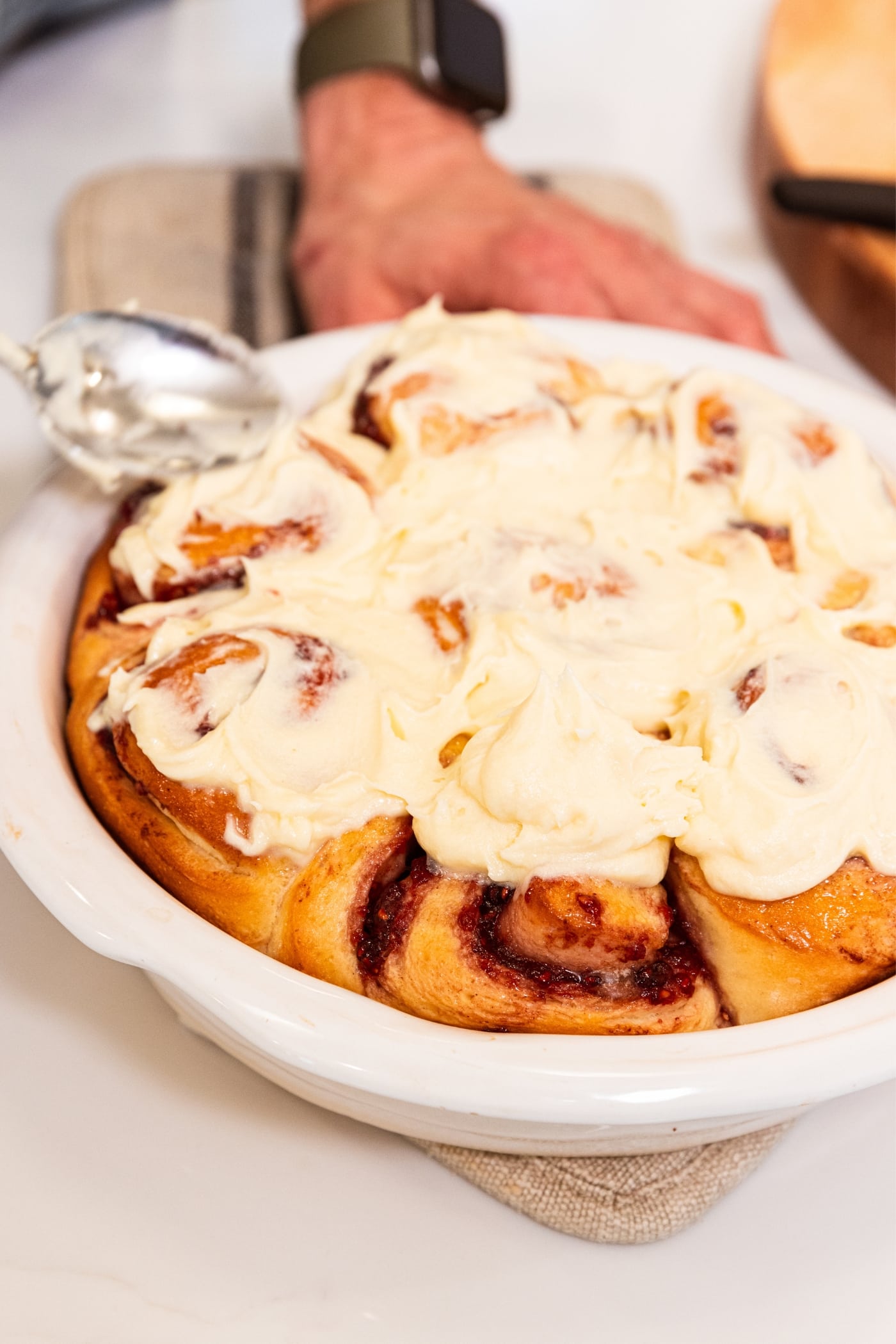 Hand using spoon to smooth cream cheese frosting over the top of raspberry cinnamon rolls in white baking dish on white surface.
