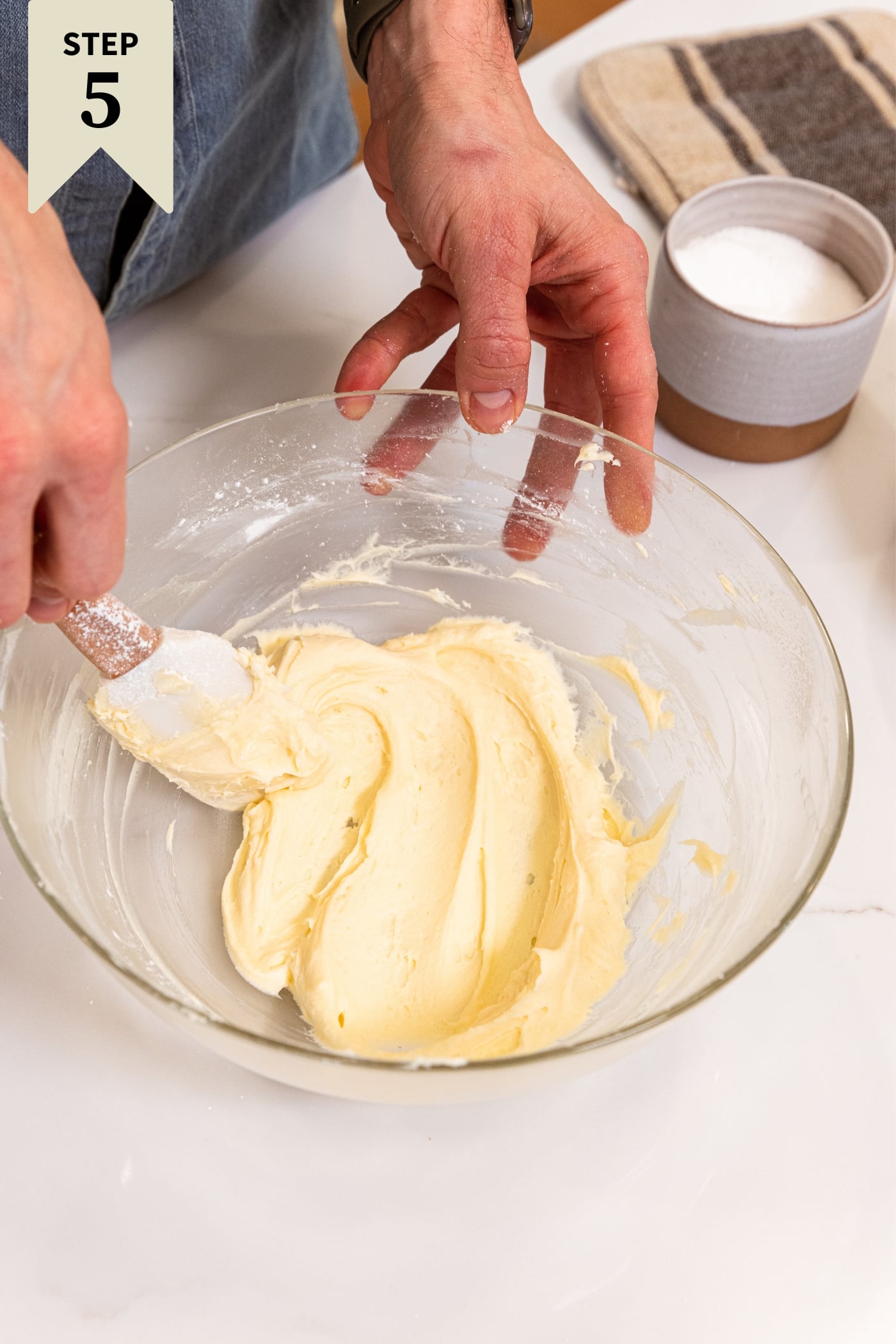 Medium glass bowl sitting on white countertop filled with soft cream cheese with hand spreading it with plastic spatula.