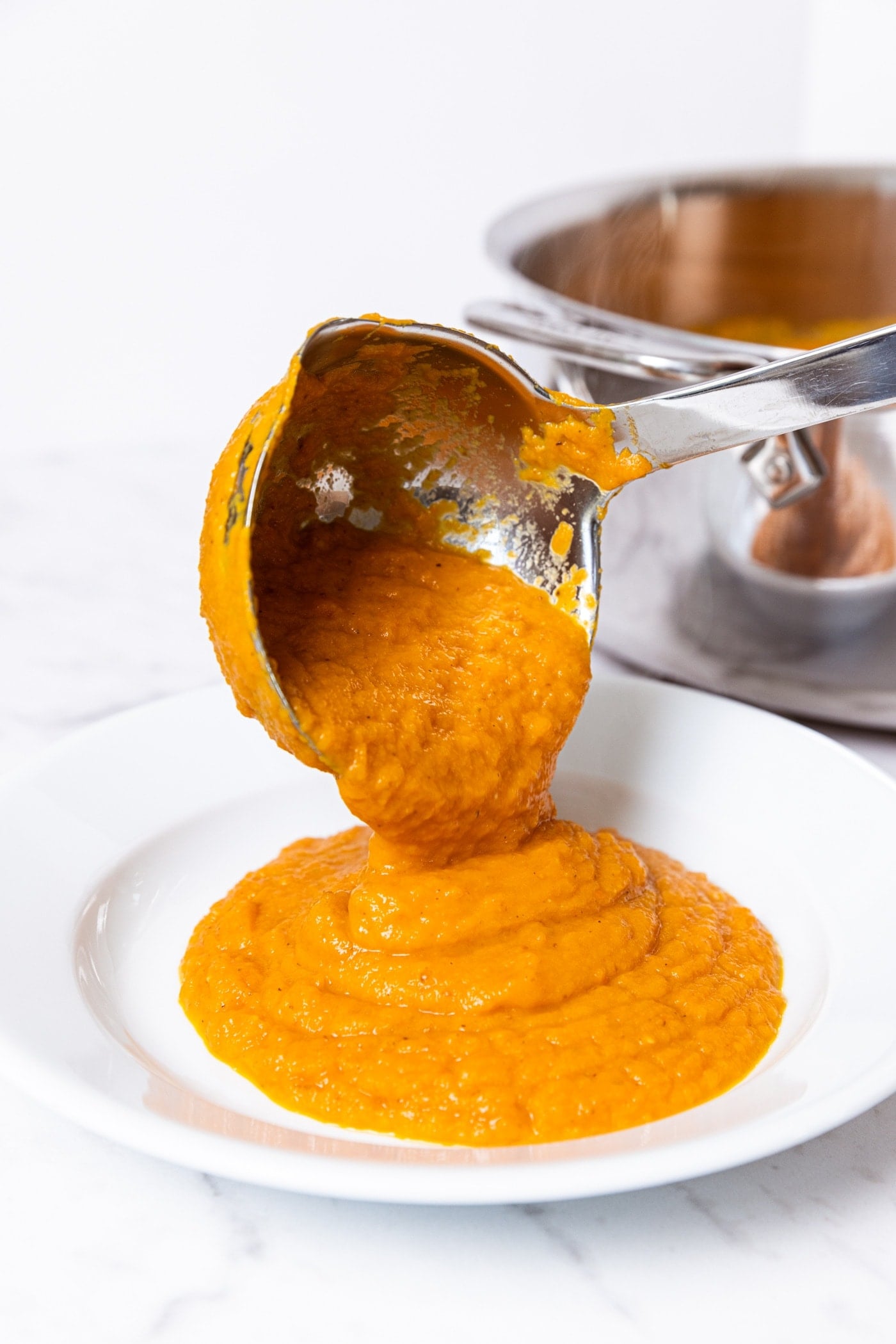 White plate bowl with ladle placing roasted carrot soup inside showing puréed texture of the soup with metal stockpot with rest of soup in background.