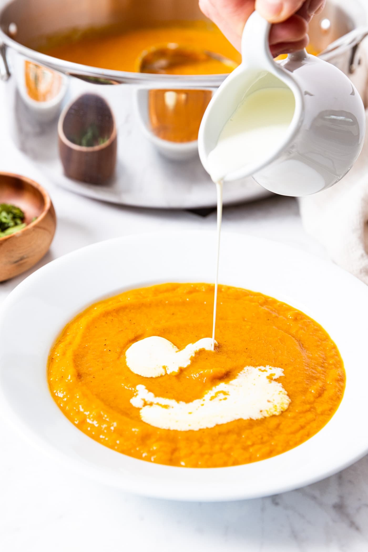 Hand holding white creamer jar pouring heavy cream on top of orange colored roasted carrot soup in large white plate bowl on marble surface with stockpot filled with rest of soup in background.