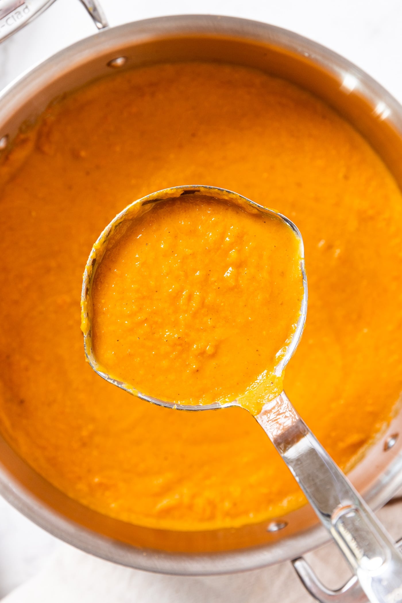 Metal stockpot filled with blended orange-colored roasted carrot soup with ladle picking up a serving showing texture.