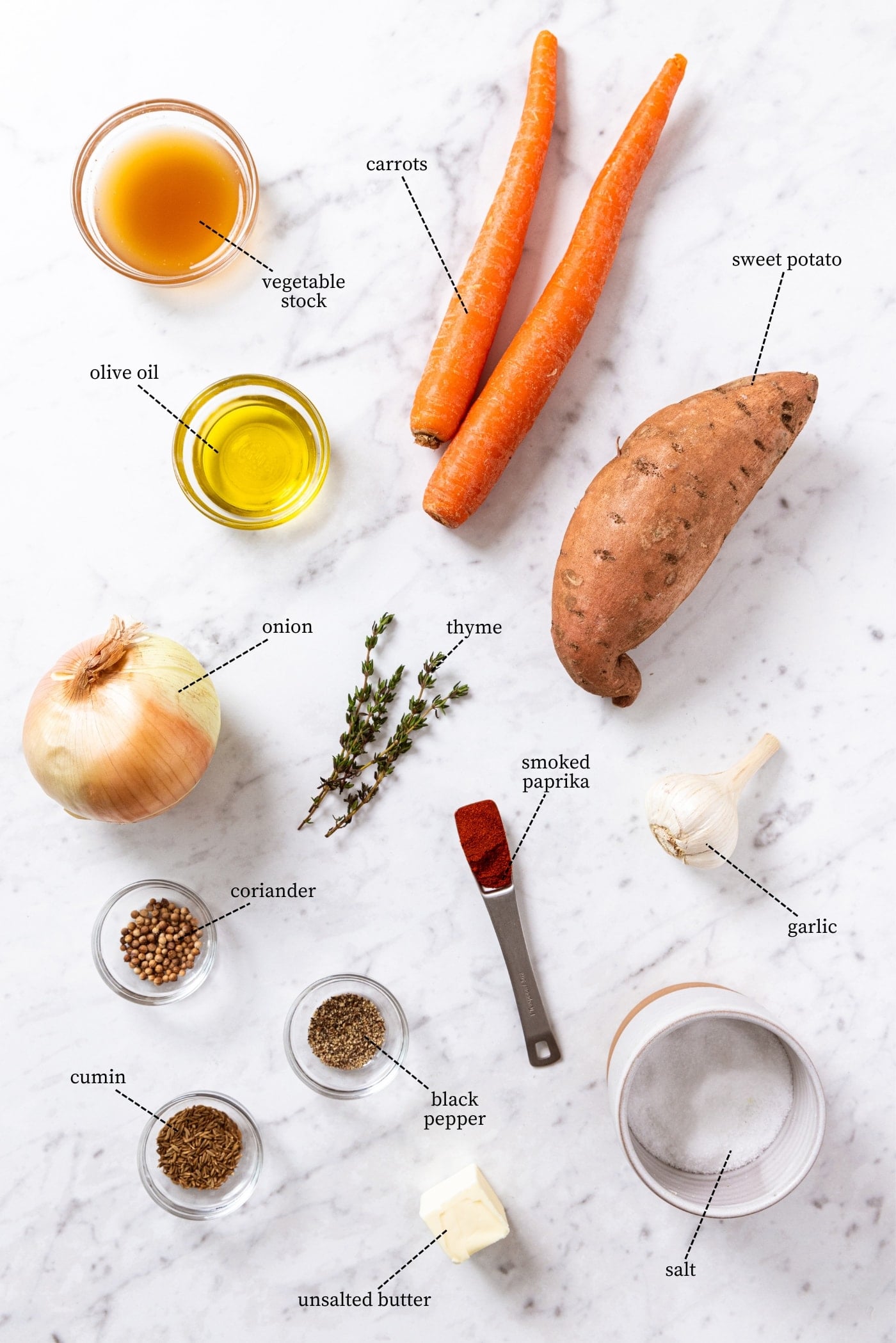 White marble surface filled with all ingredients needed to prepare a roasted carrot soup including carrots, sweet potato, onion, herbs, pieces, stock, olive oil, and more.