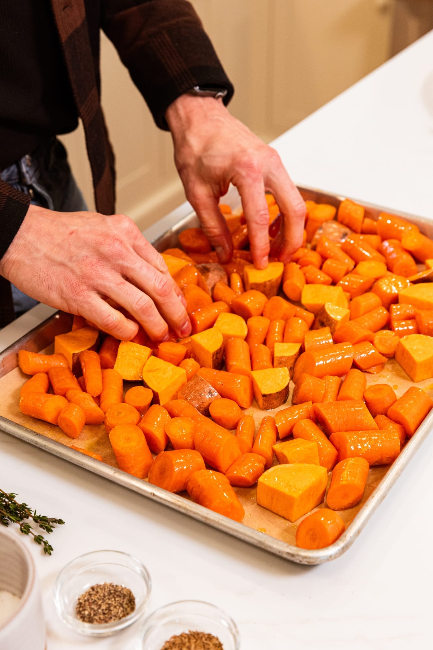 Two hands arranging sliced orange carrots and sweet potatoes on a parchment-lined baking sheet with olive oil and salt and black pepper on top all on white surface in kitchen.