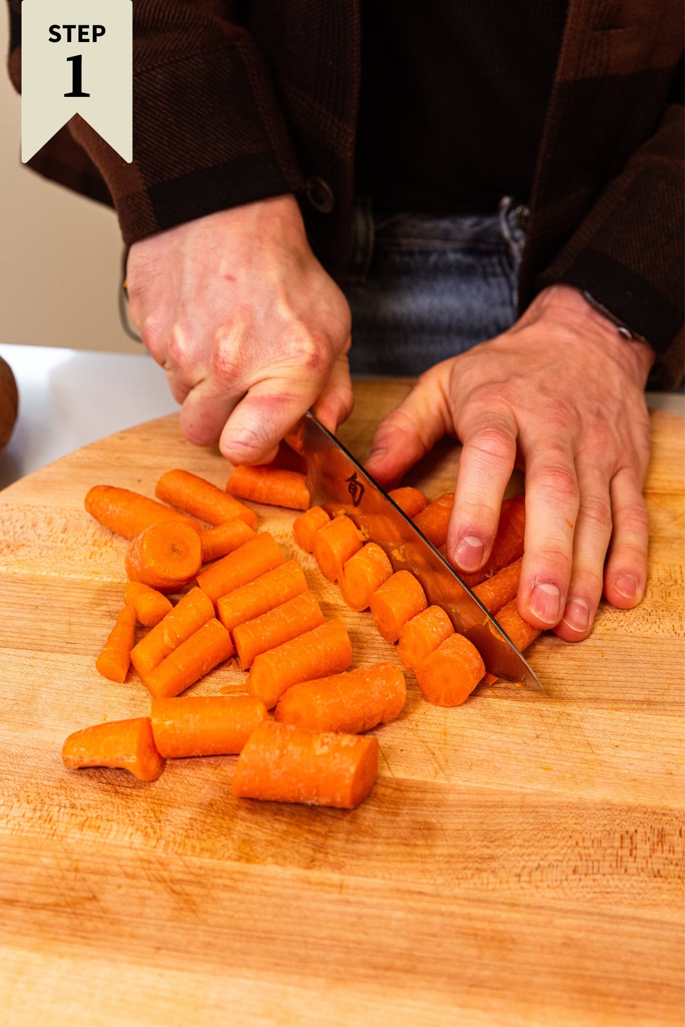 Hand using large knife to chop orange carrots into chunks on wood cutting board.