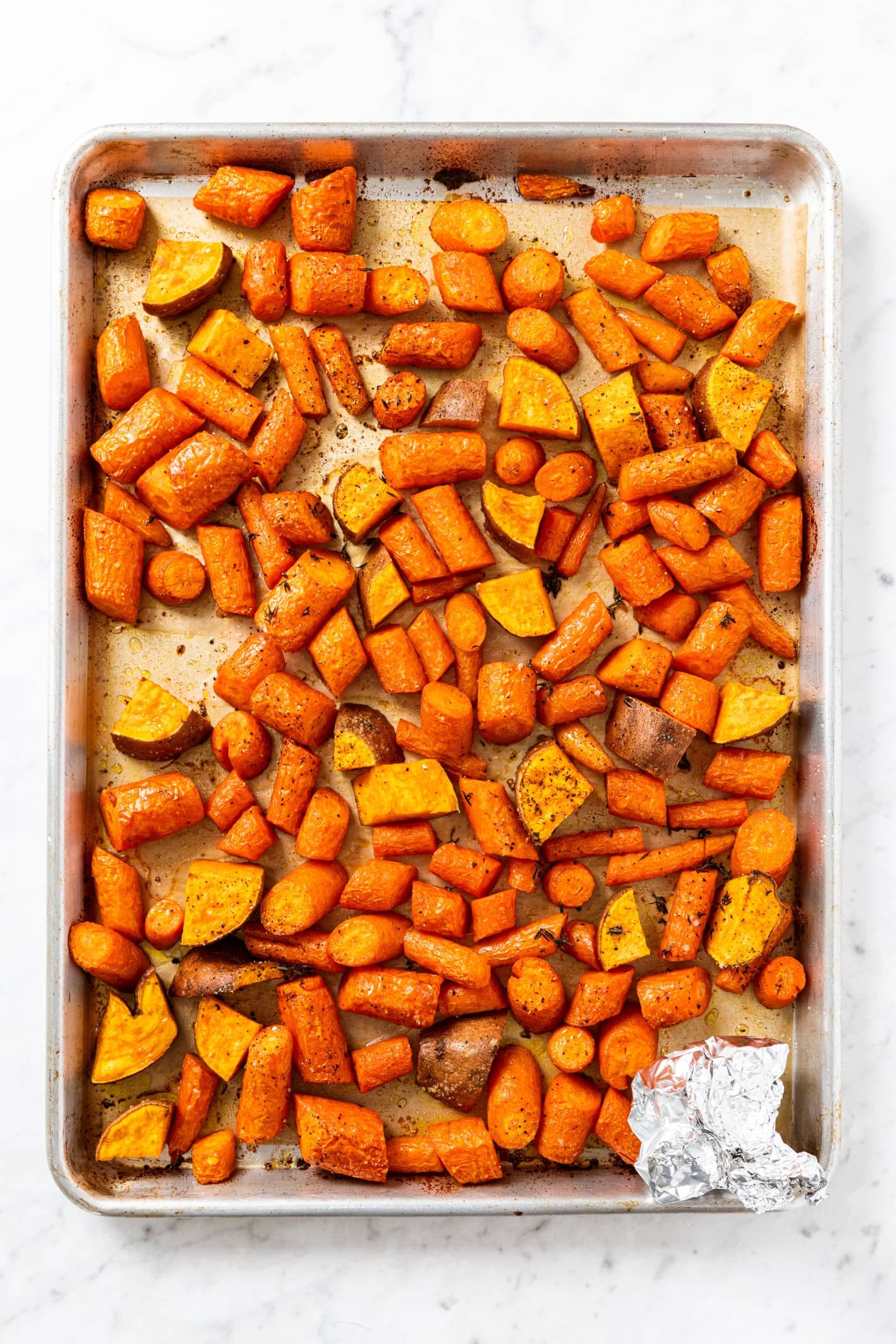 Metal baking sheet with piece of parchment paper with sliced and baked pieces of sweet potato and carrot right after coming out of the oven.