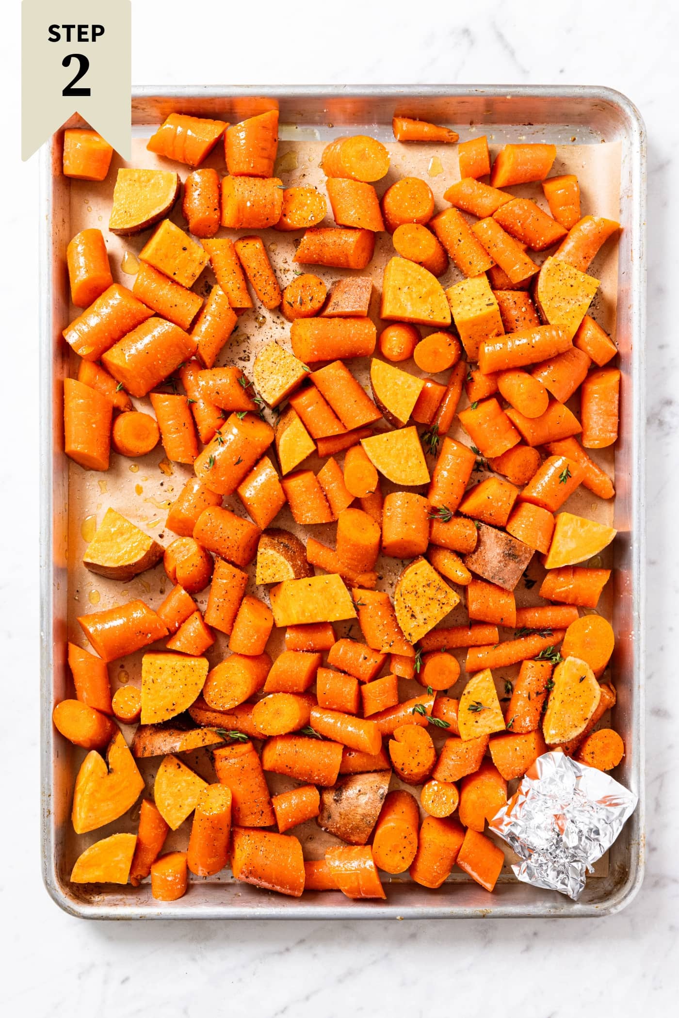Large metal baking sheet with piece of parchment with unbaked sliced carrots and sweet potato and small tin foil packet sitting on marble surface right before going into the oven.