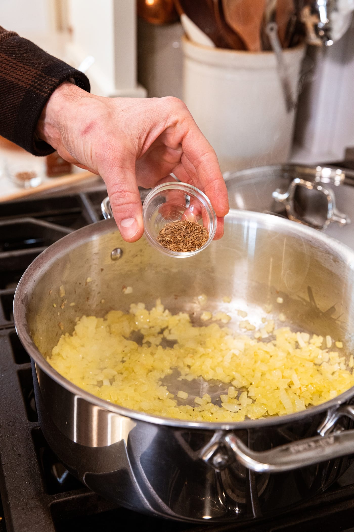 Hand holding glass bowl filled with spices about to pour the spices into a large metal stockpot filled with lightly browned onion cooked in melted butter.
