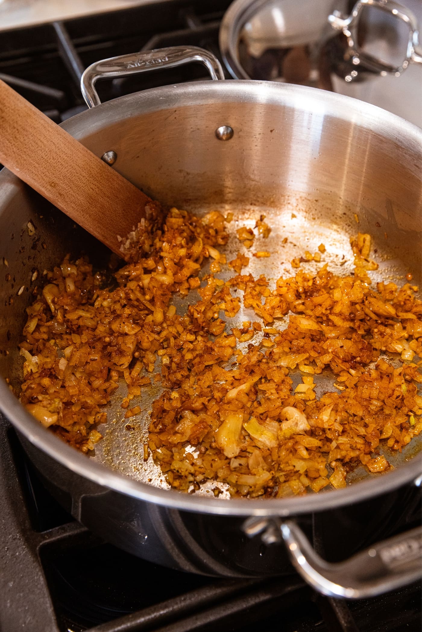 Wooden spatula resting on the bottom of a large metal stockpot filled with browned onion and spices in melted butter on stovetop.