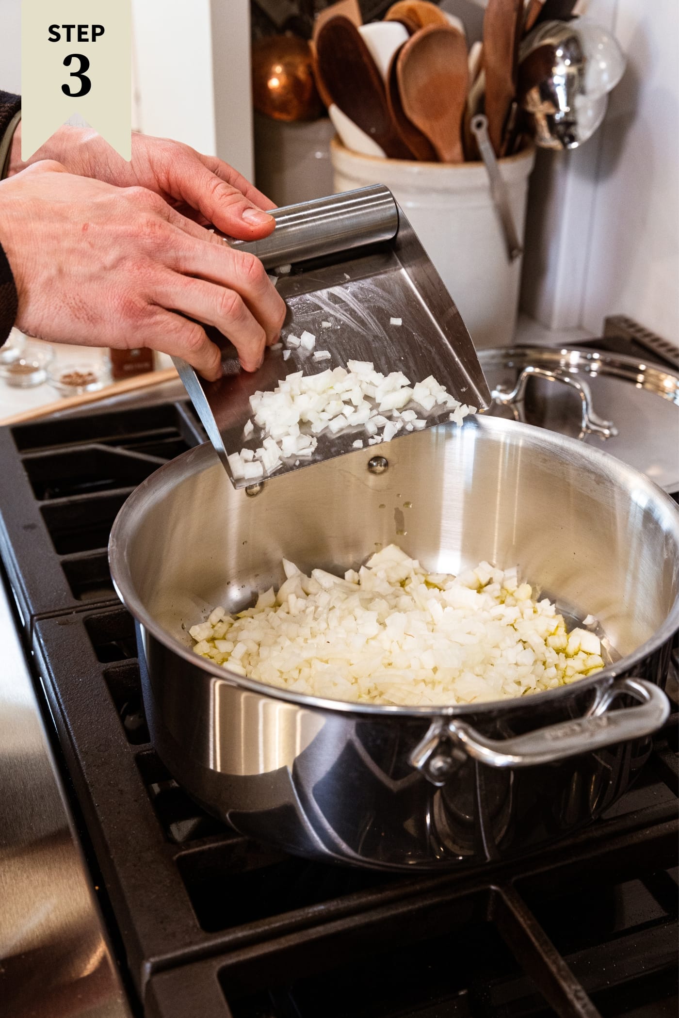 Hand using metal scoop to push chopped onion into a large metal stockpot on the stovetop with melted butter in the bottom.