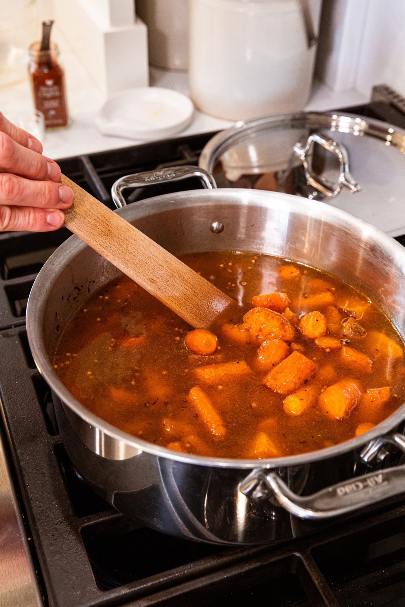 Hand using wooden spatula to stir soup in large stockpot on stovetop with roasted carrots floating on the top.