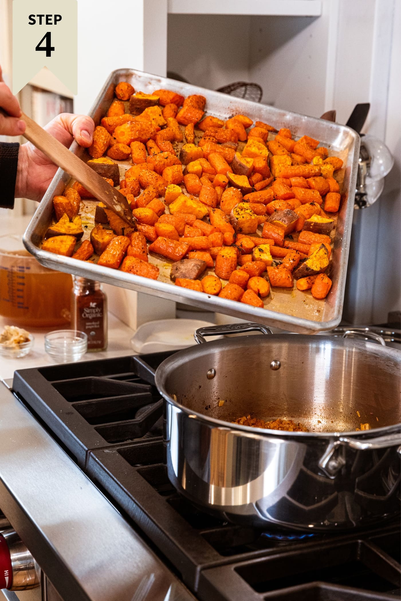 Hand using wooden spatula to push roasted pieces of carrot and sweet potato from sheet pan into large metal stockpot on the stovetop.