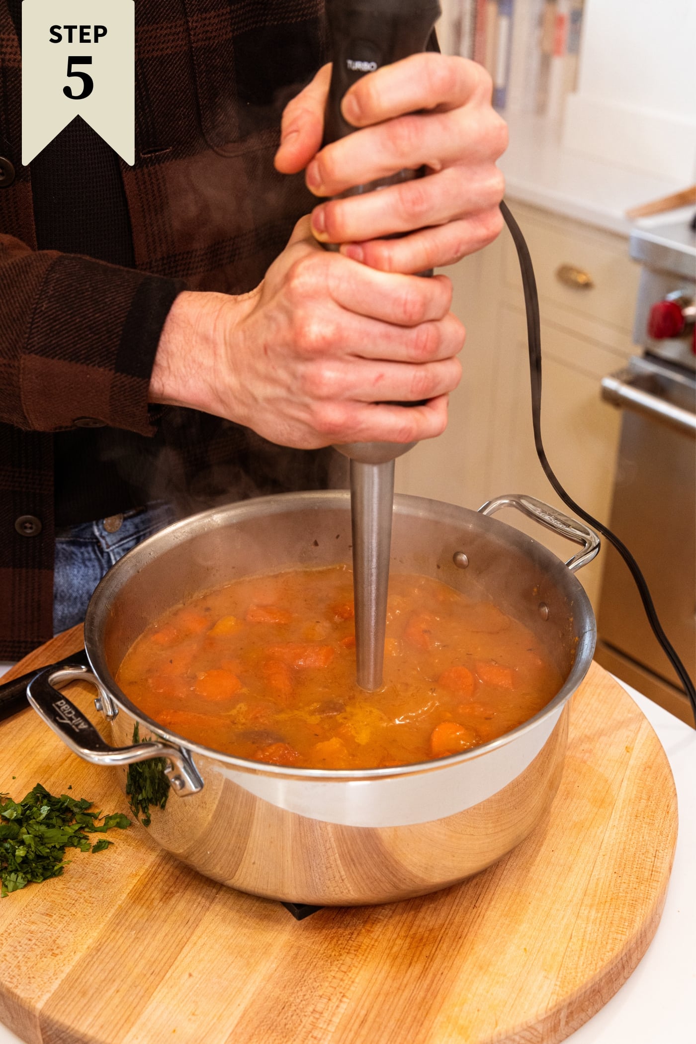 Two hands holding onto immersion blender blending together roasted carrot soup in large metal stockpot sitting on wood cutting board in kitchen.