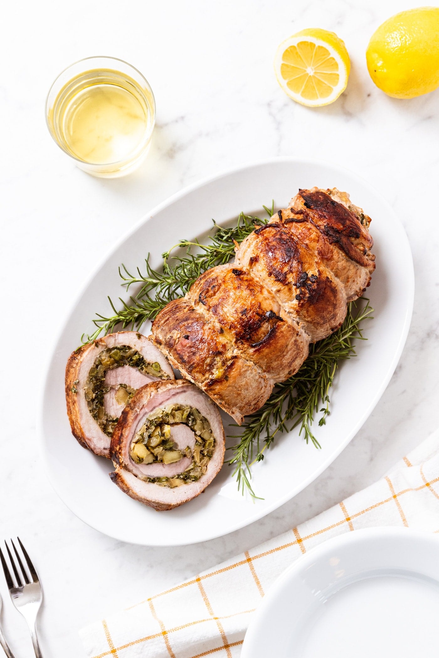 White marble surface with white platter with pork roulade sitting on a bed of fresh herbs with two slices on the side of the plate with glass of white wine and sliced lemon to the side.