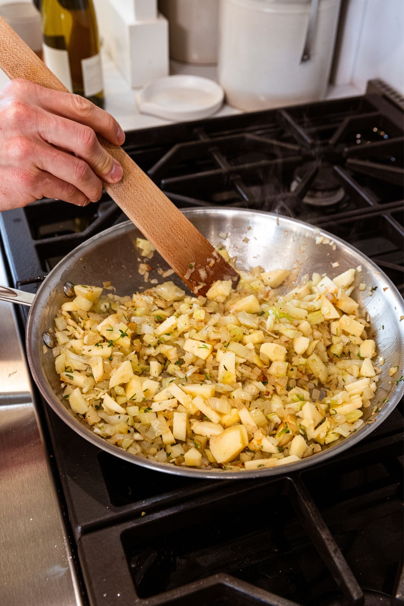 Hand holding wooden spatula mixing together browned onion and fennel mixture with herbs all in metal skillet on stovetop.
