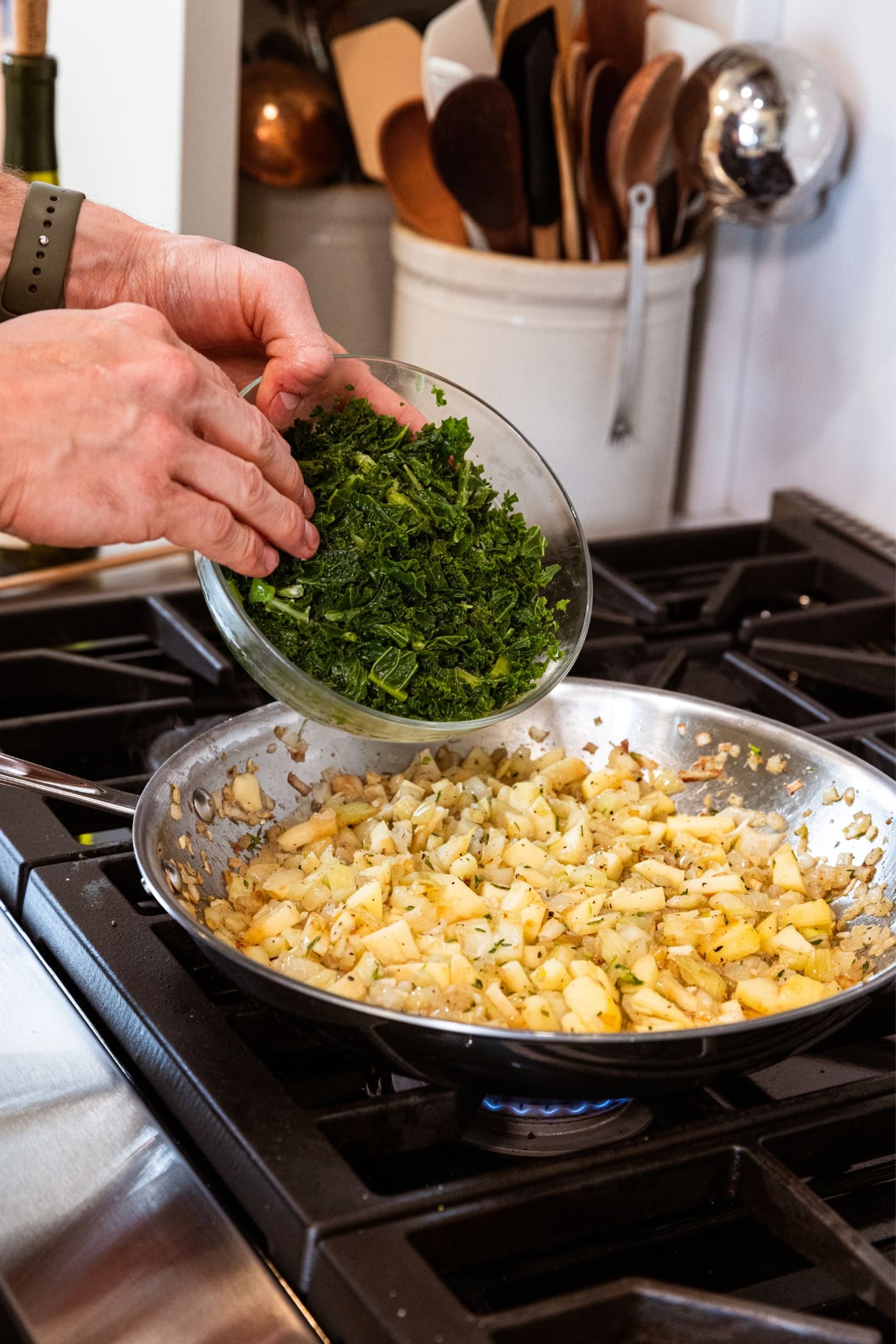Hand holding glass bowl about to push frozen kale into a metal skillet on the stovetop filled with browned onion and fennel mixture.