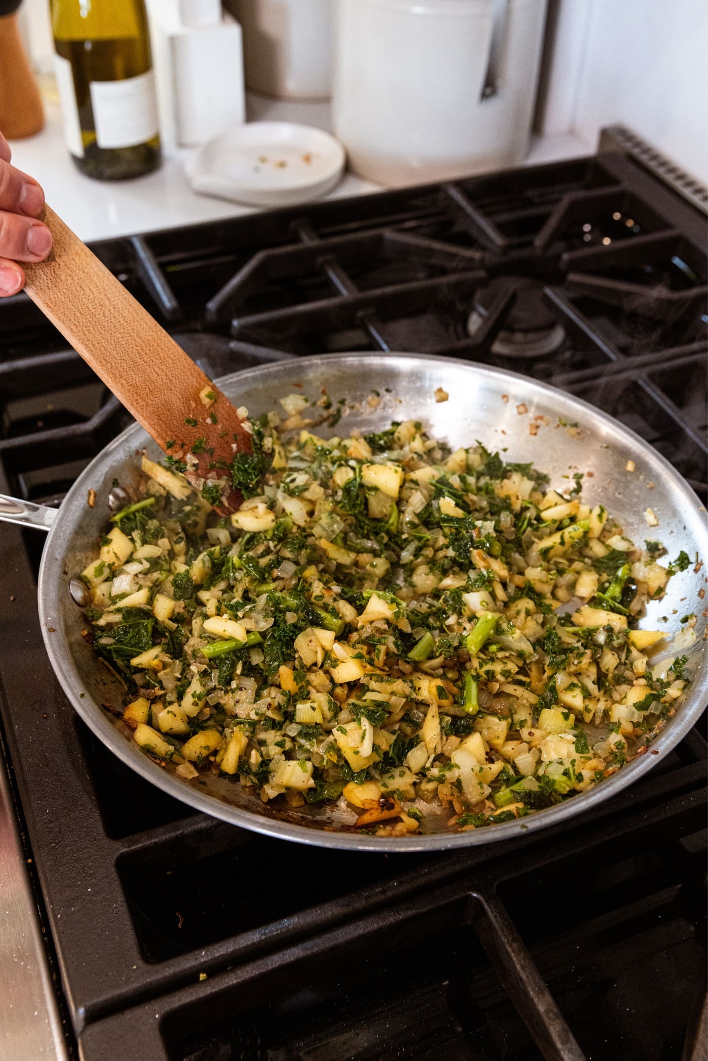 Hand holding wooden spatula mixing together cooked onion and fennel with wilted kale all in metal skillet on stovetop.