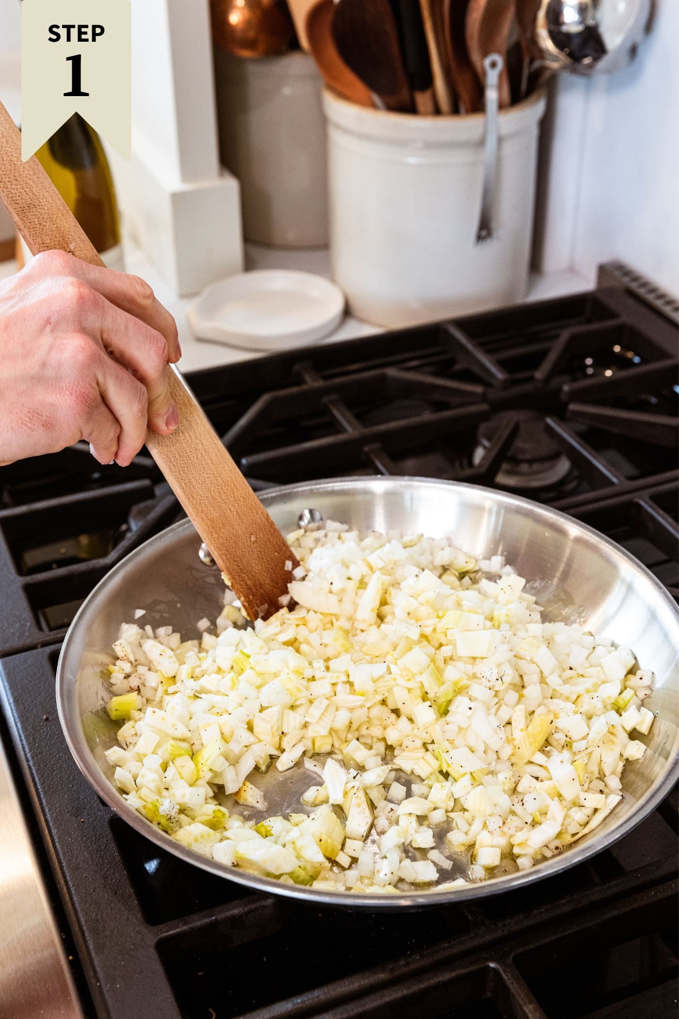 Hand holding wooden spatula mixing together onion and fennel in a metal skillet on the stovetop.