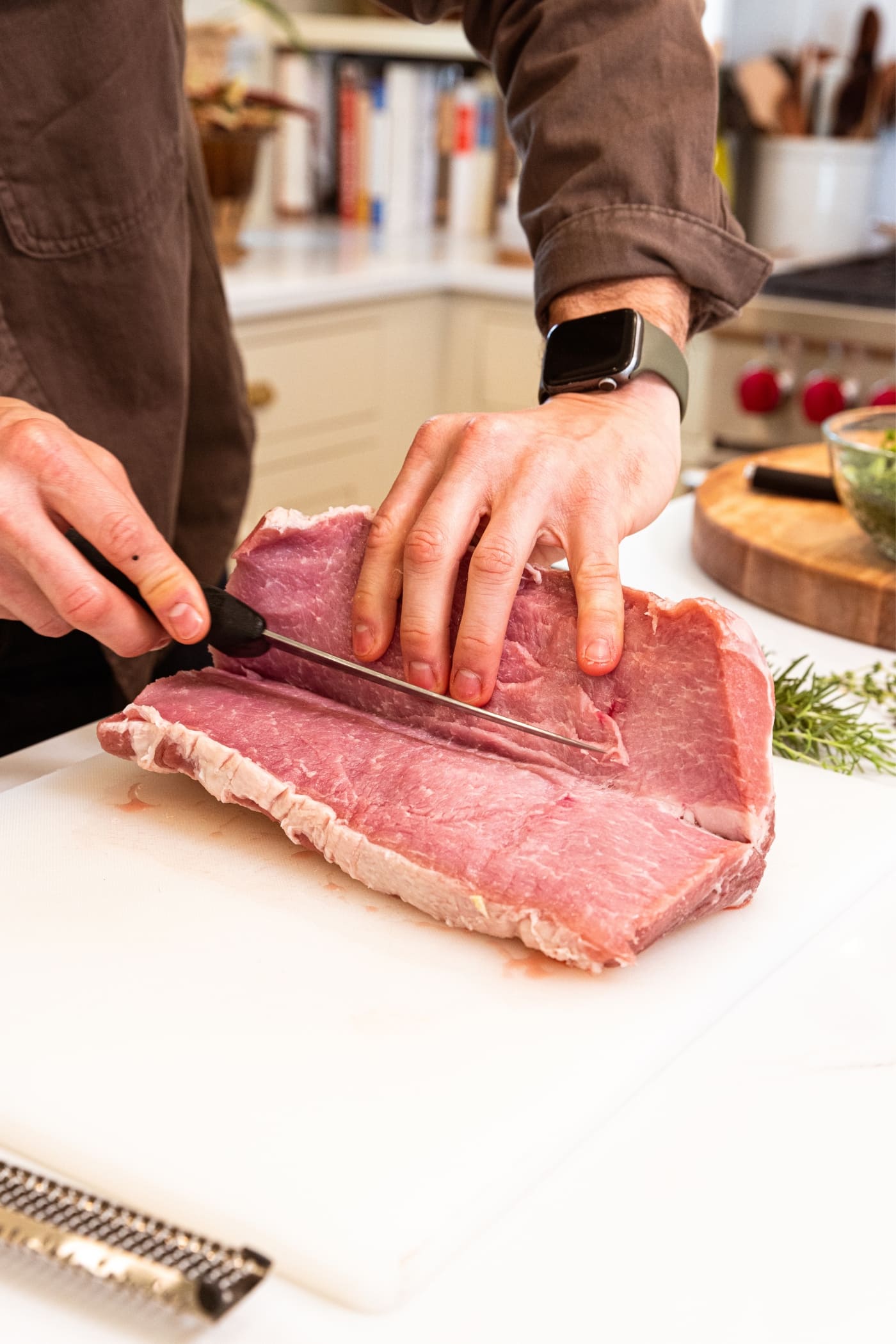 Two hands using knife to slice open a pork loin on a white cutting board with herbs in background.