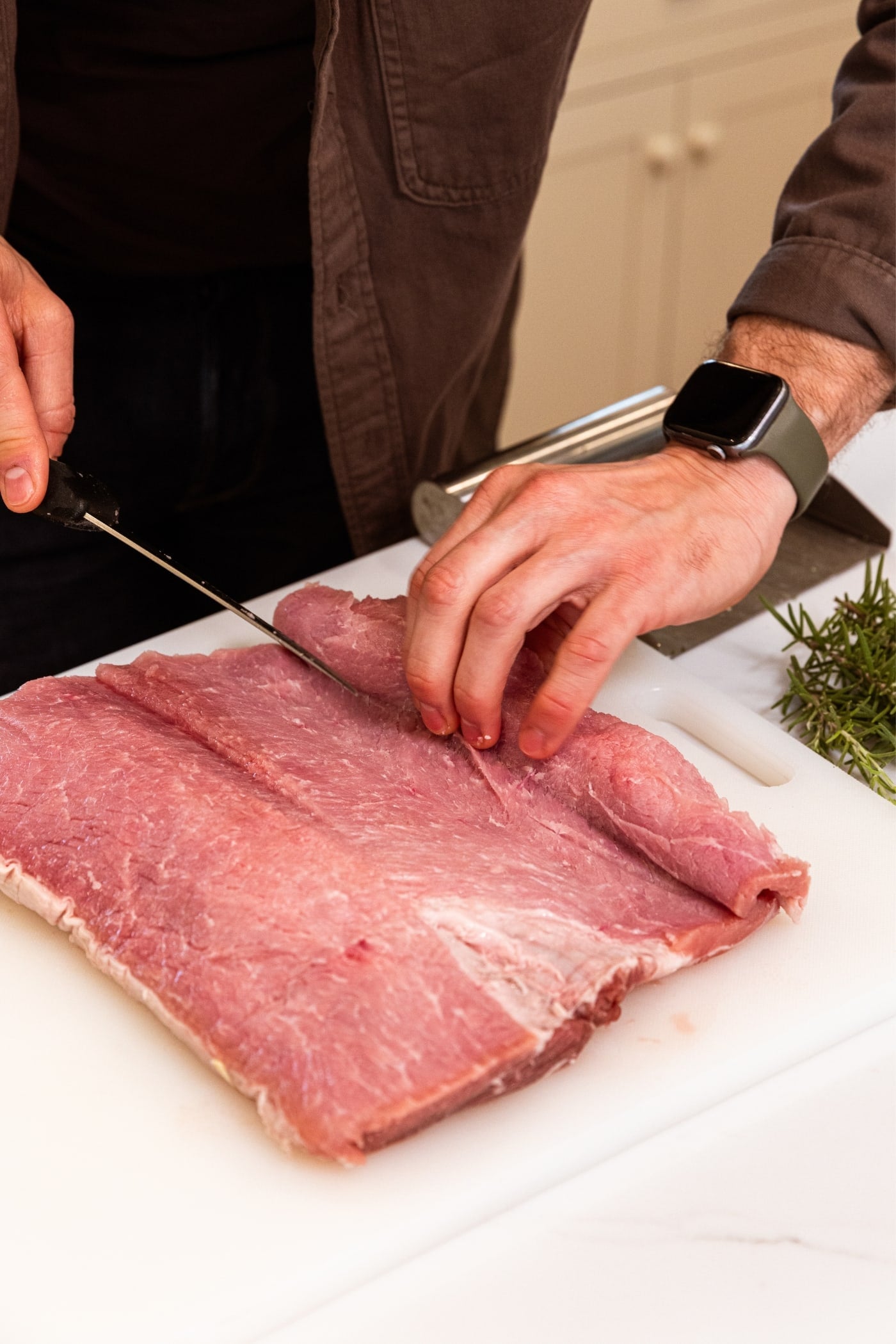 Two hands using knife to butterfly pork loin on white cutting board with fresh herbs in background.