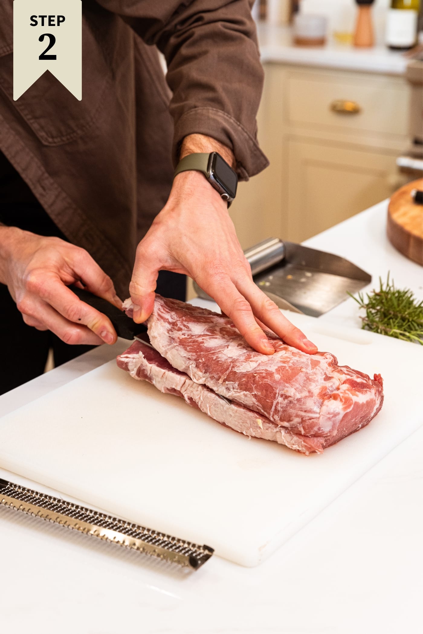 Hands using knife to cut pork roast in half on white cutting board with fresh herbs in the background.