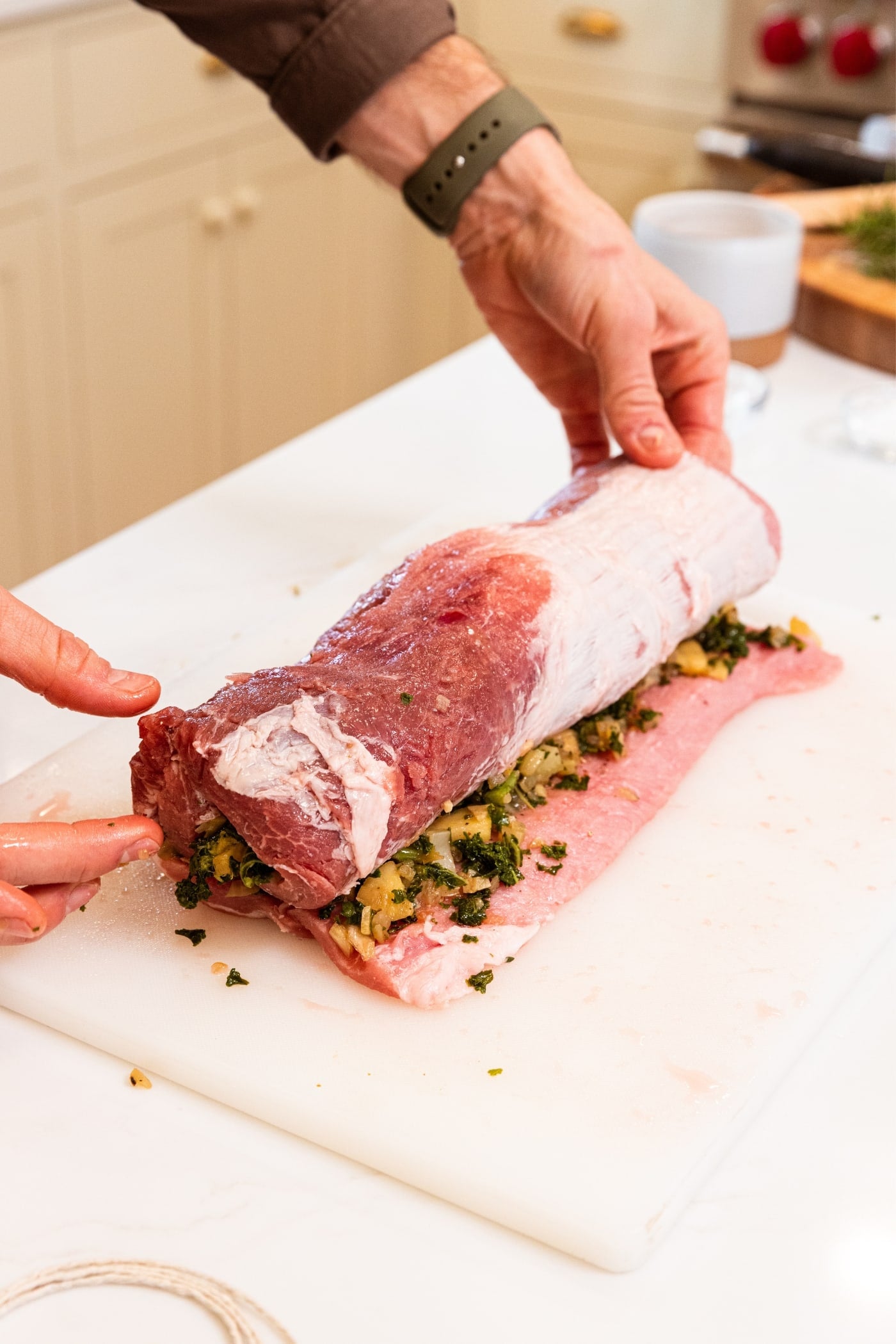 Two hands rolling up raw pork loin with cooked filling in the middle on white cutting board.