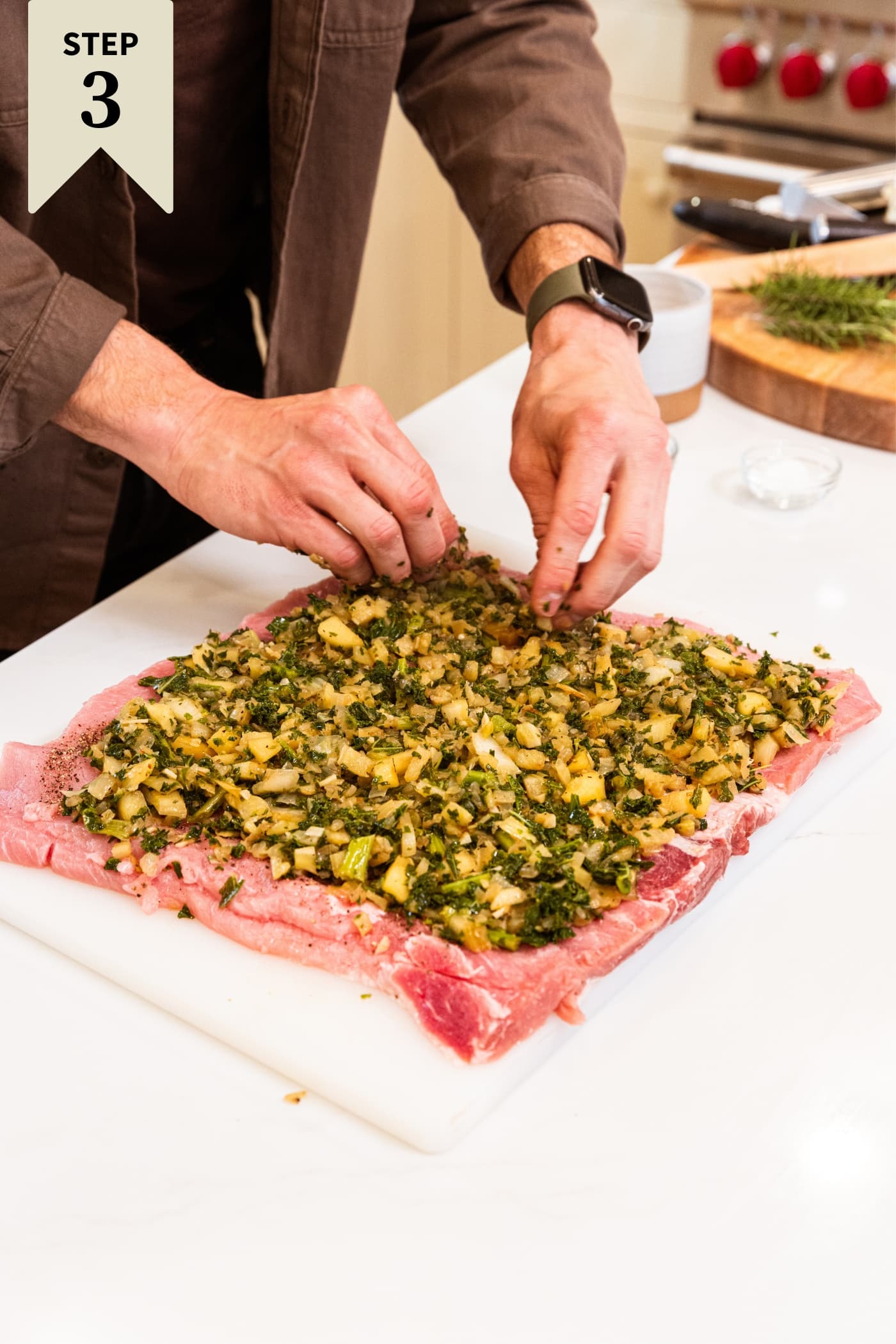 Hands leveling out onion, kale, and fennel mixture ton top of raw pork loin sitting on white cutting board.