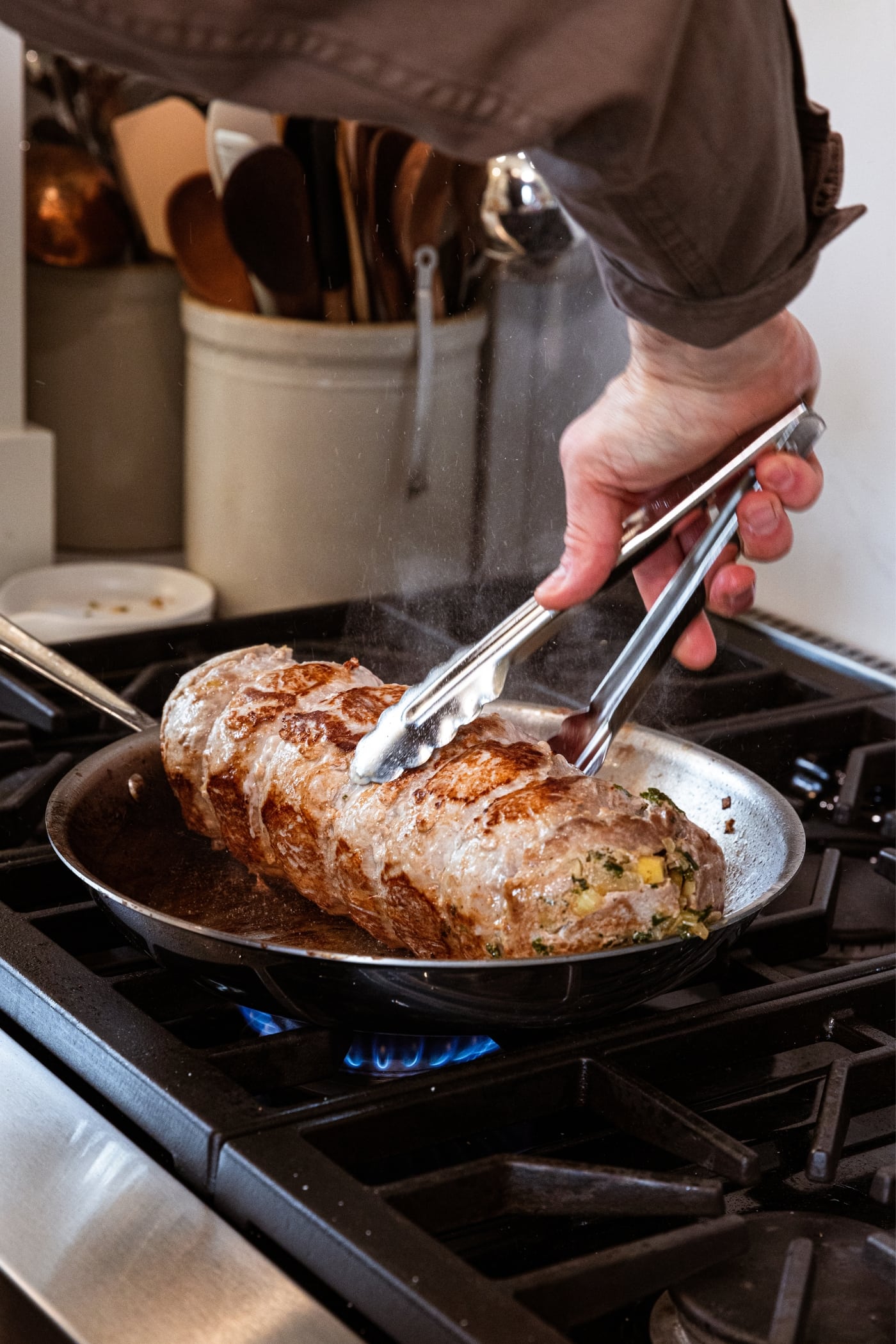 Hand using tongs to turn pork roulade in metal skillet on stovetop with browned sides.