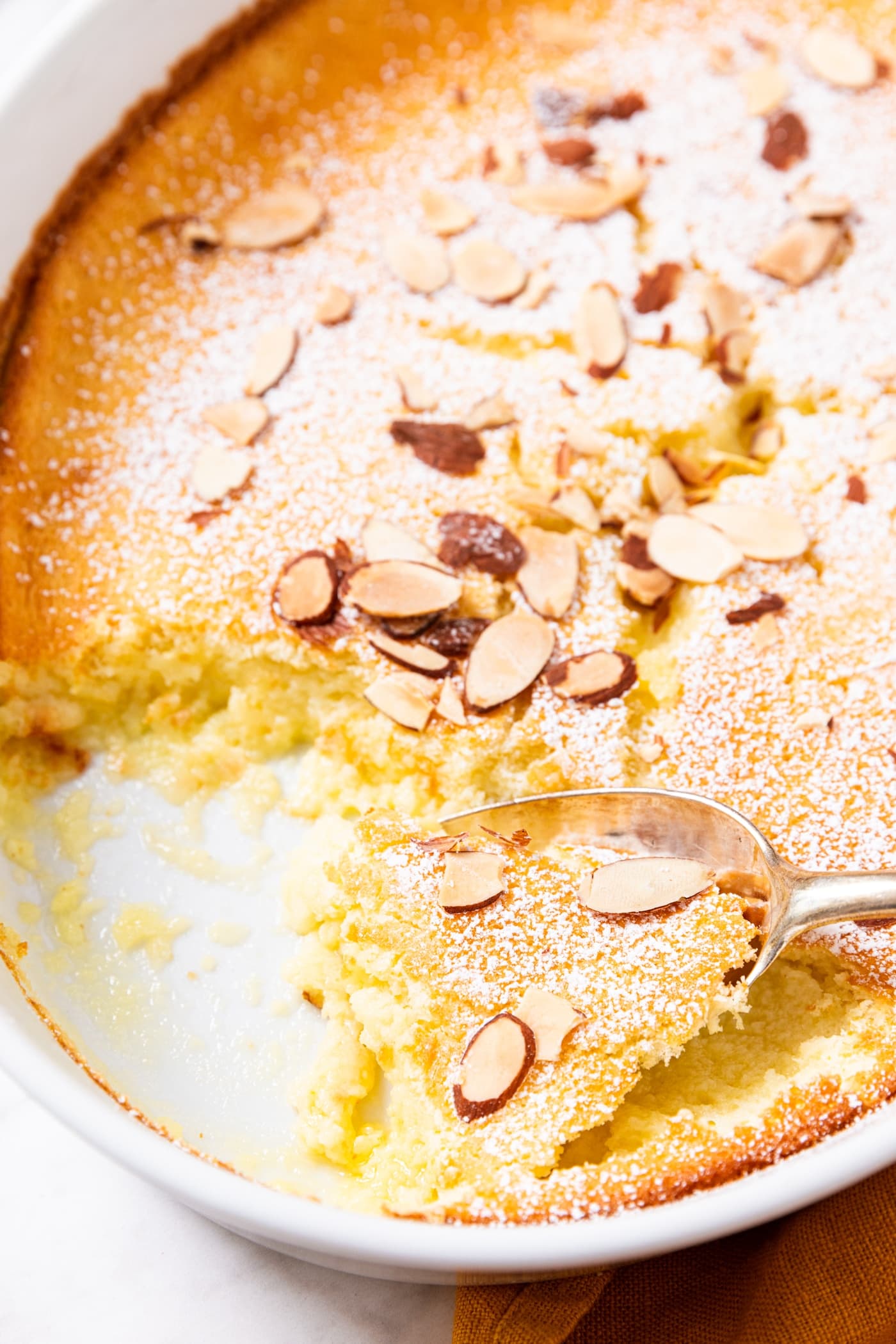 Scoop of lemon pudding cake resting on a spoon in an oval baking dish with baked cake in it topped with powdered sugar and toasted almonds with a few servings already removed.