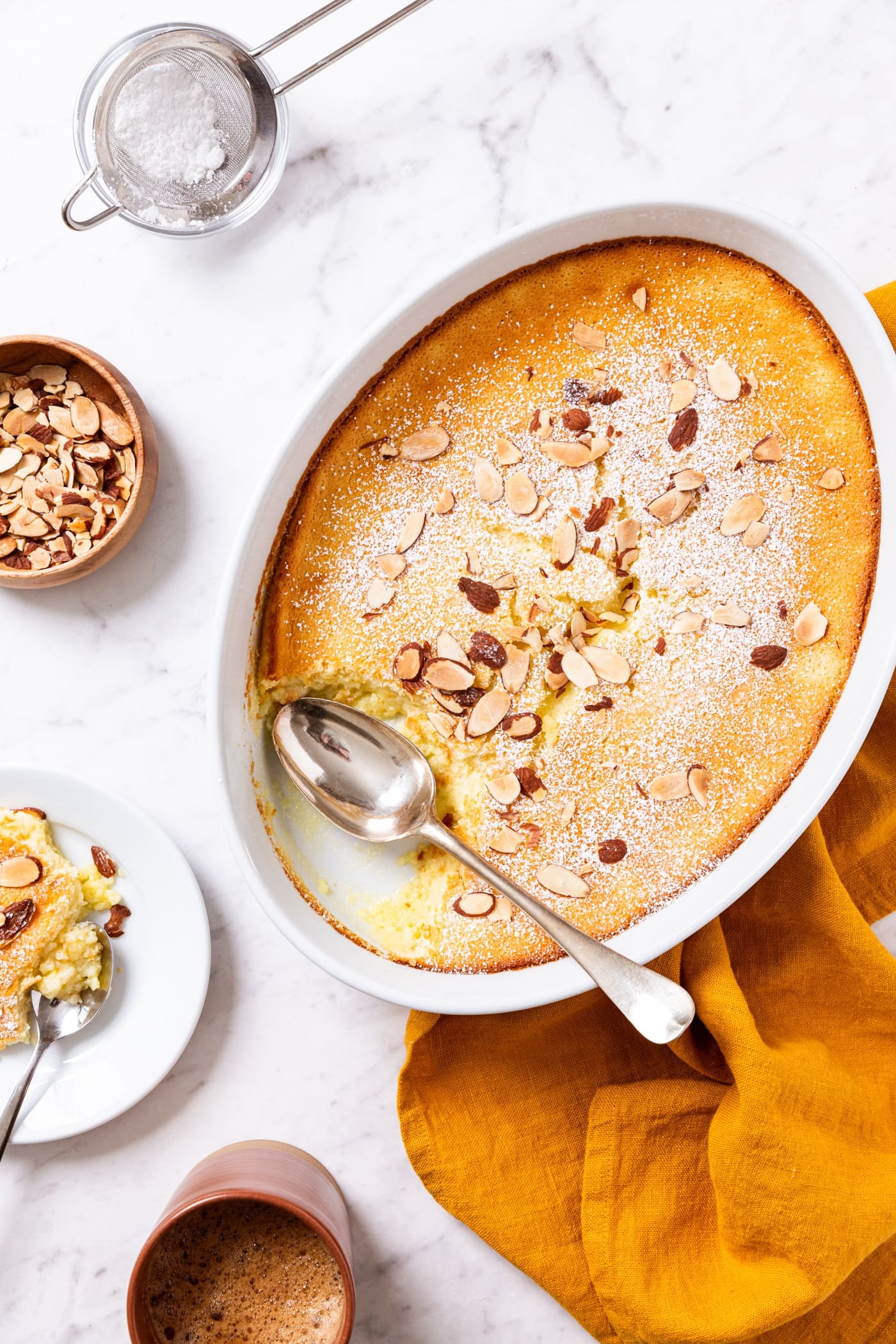 White marble surface with oval serving dish with baked lemon pudding cake dusted with powdered sugar and toasted almonds with a serving sitting on a small white plate with a cup of coffee to the side.