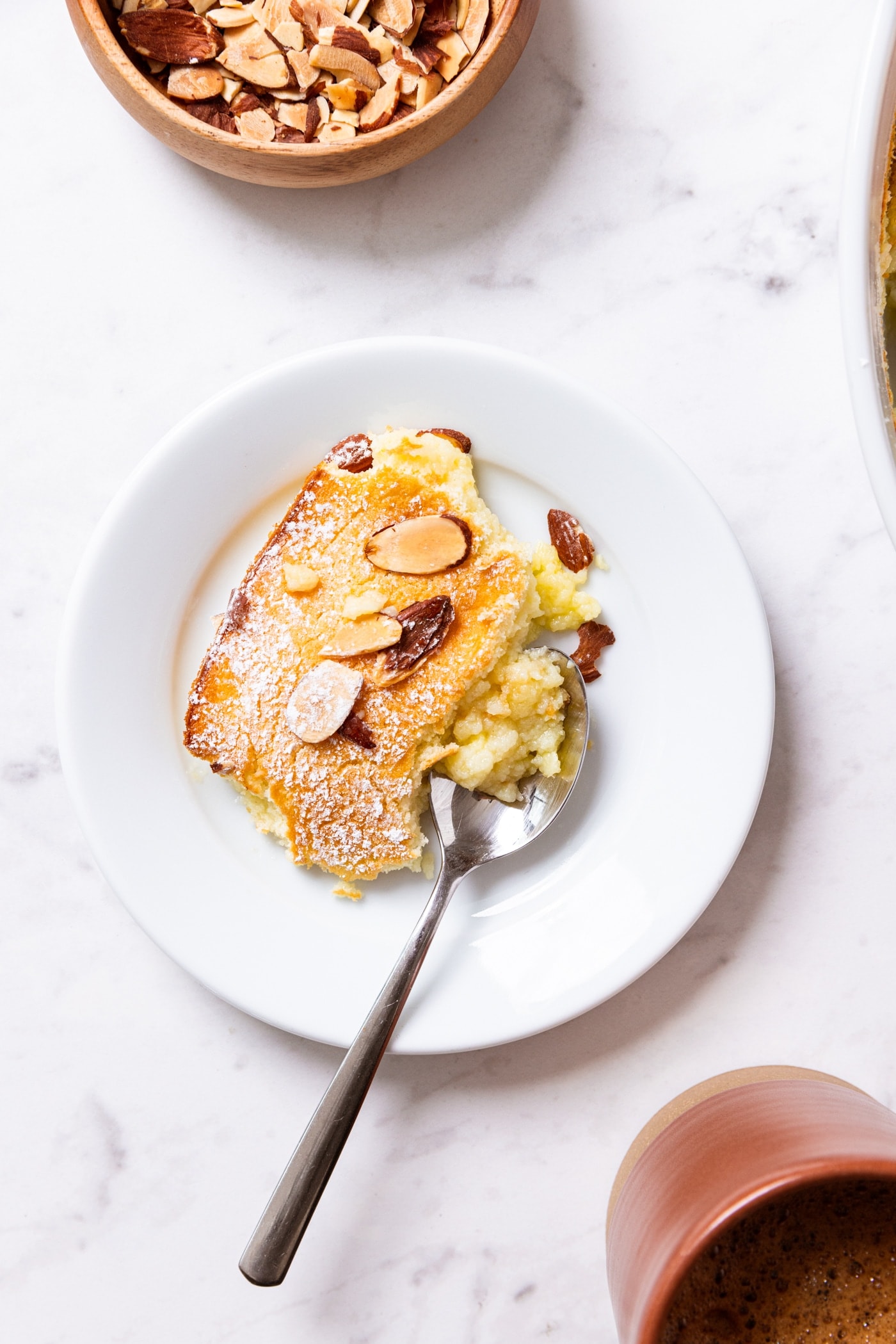 White marble surface with white plate with spoon to the side with serving of lemon pudding cake with sliced almonds on top with cup of coffee to the side.