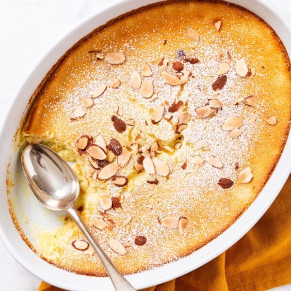 Oval baking dish sitting on marble surface filled with baked lemon pudding cake dusted with powdered sugar and topped with toasted almonds with spoon resting on side of platter after removing a serving.
