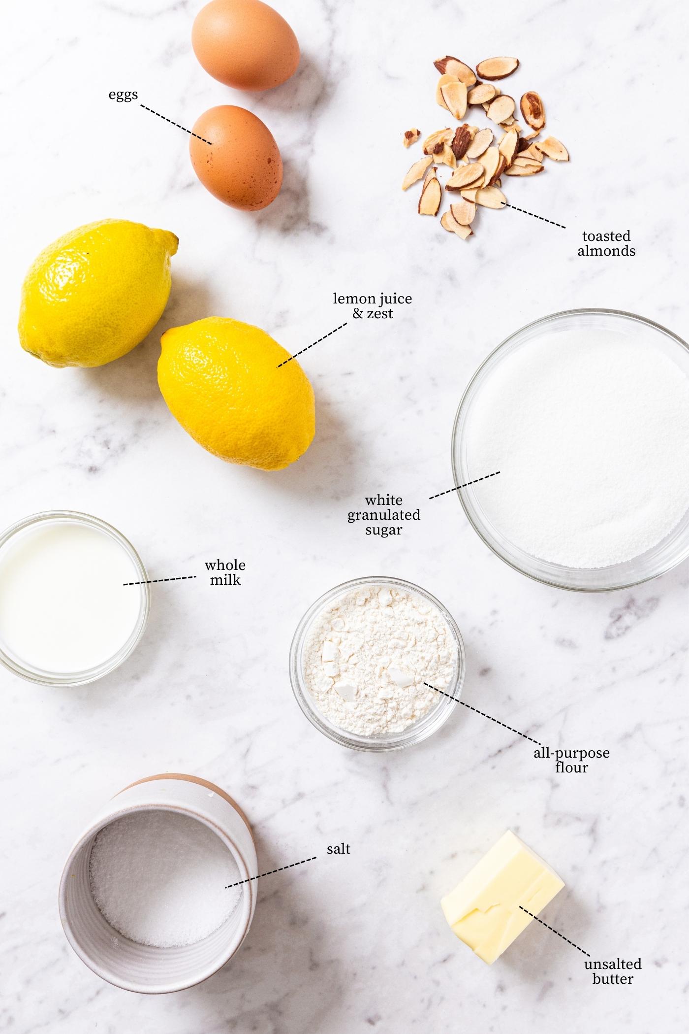 White marble surface filled with all ingredients needed to make a lemon pudding cake including lemons, eggs, flour, sugar, slat, butter, and more.