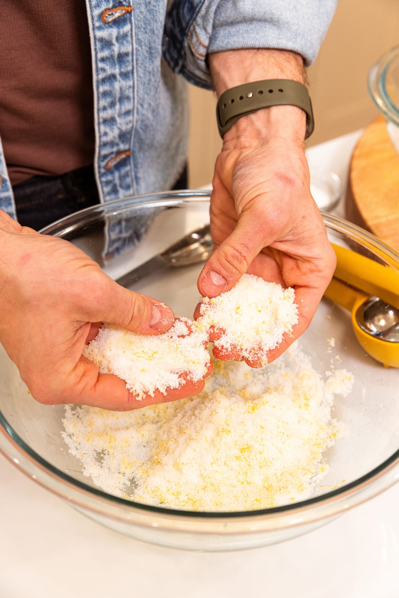 Two hands holding sugar mixing together with lemon zest in large glass bowl on white surface with wood cutting board to the side.