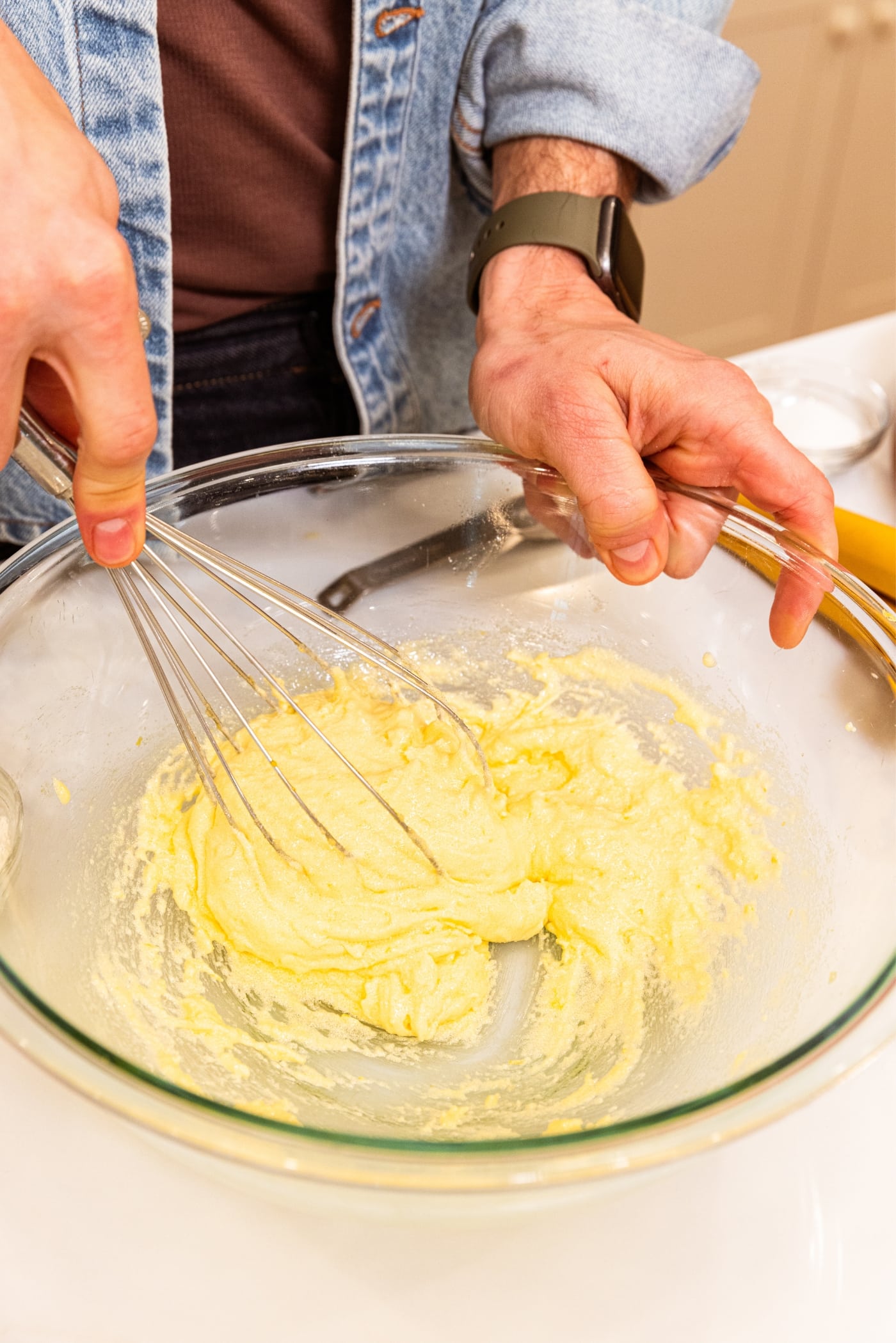 Hand holding whisk mixing everything together in a large glass bowl with light pale yellow colored batter all on white surface.