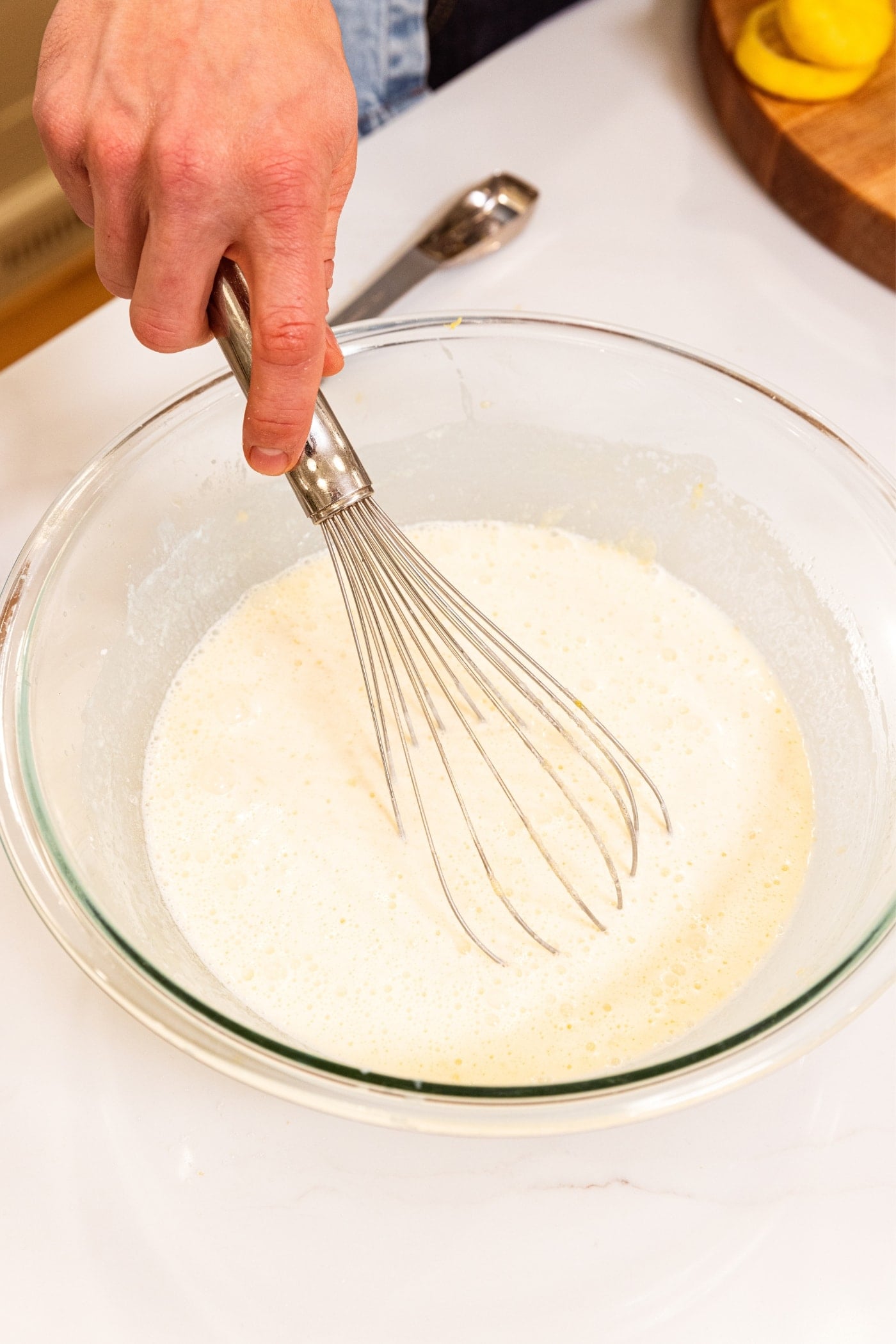 Hand holding glass bowl filled with milk-looking mixture with whisk mixing everything together on white surface.