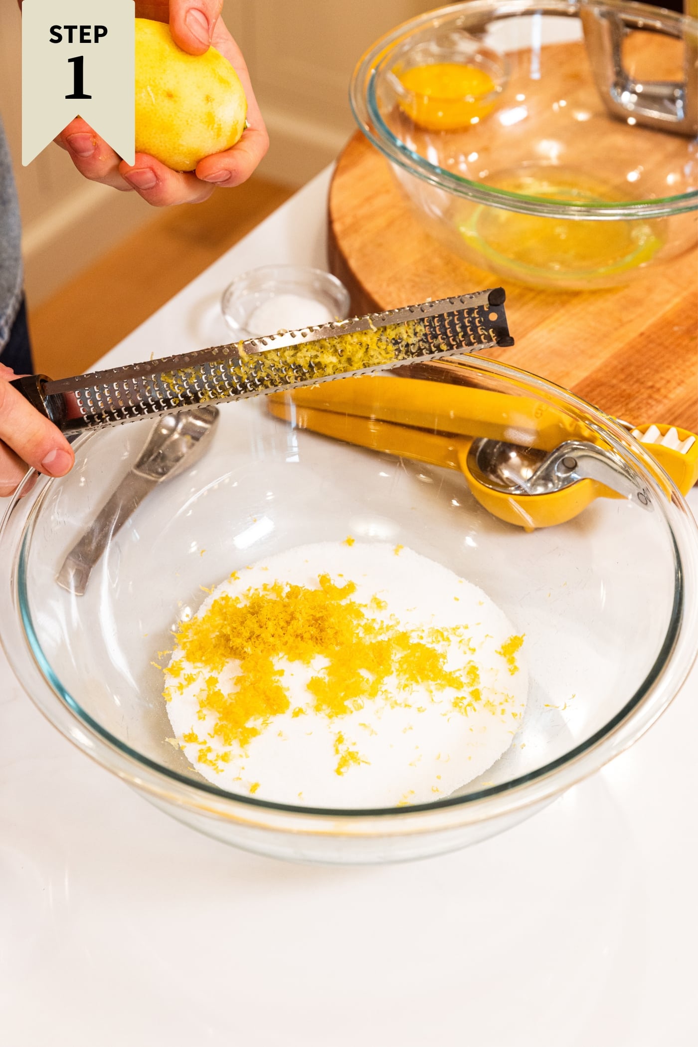 Hand holding Microplane knocking off the zested lemon zest from a lemon into a glass bowl filled with sugar and a wood cutting board in the background.