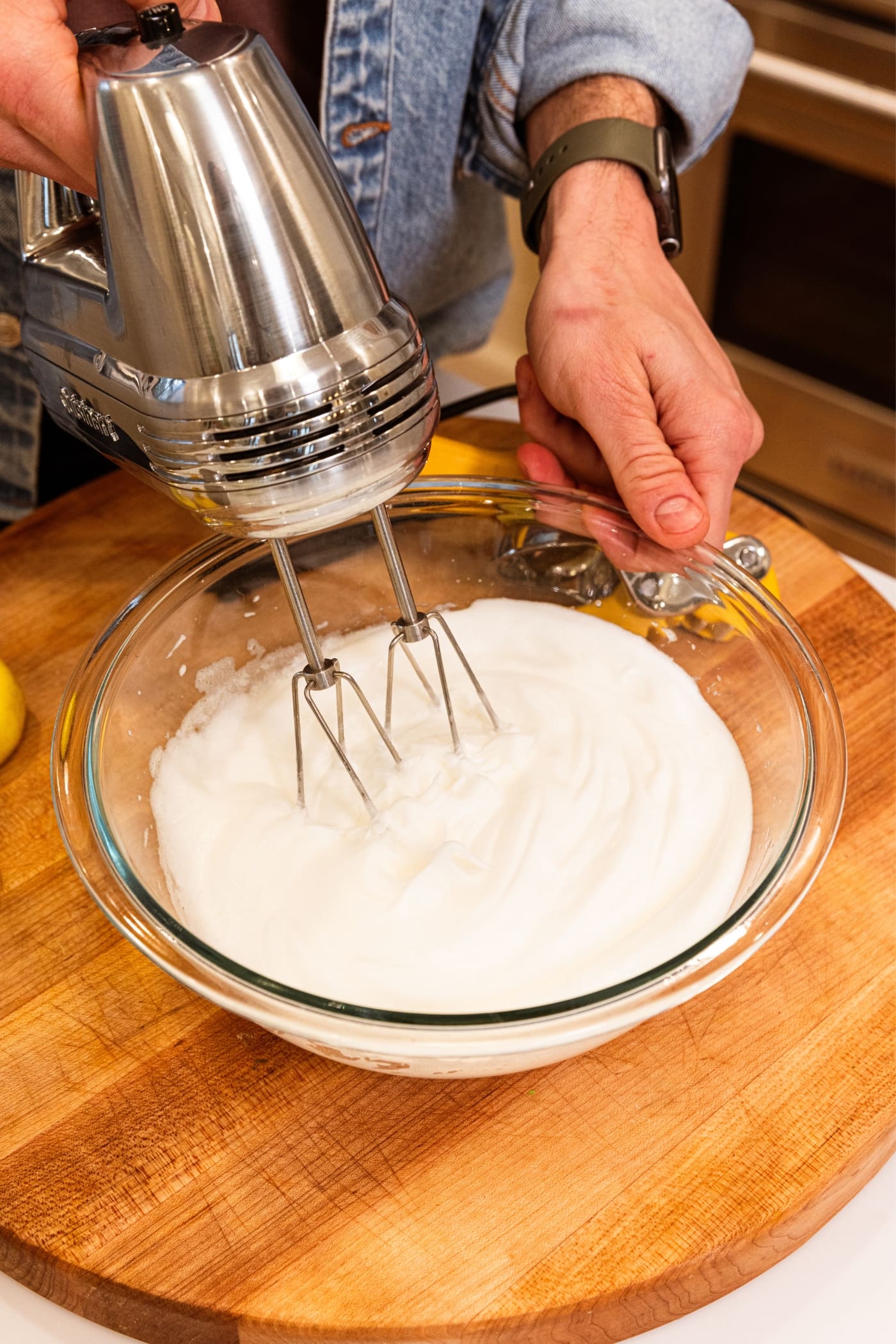 Hand holding onto the side of a glass mixing bowl filled with beaten and fluffy egg whites with hand mixer mixing everything together on wood cutting board.