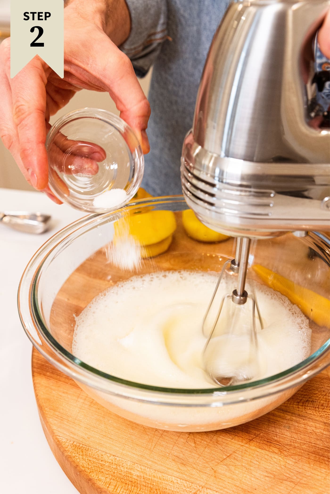 Hand pouring sugar into a glass bowl with egg whites being whipped from a small glass bowl sitting on wood cutting board.
