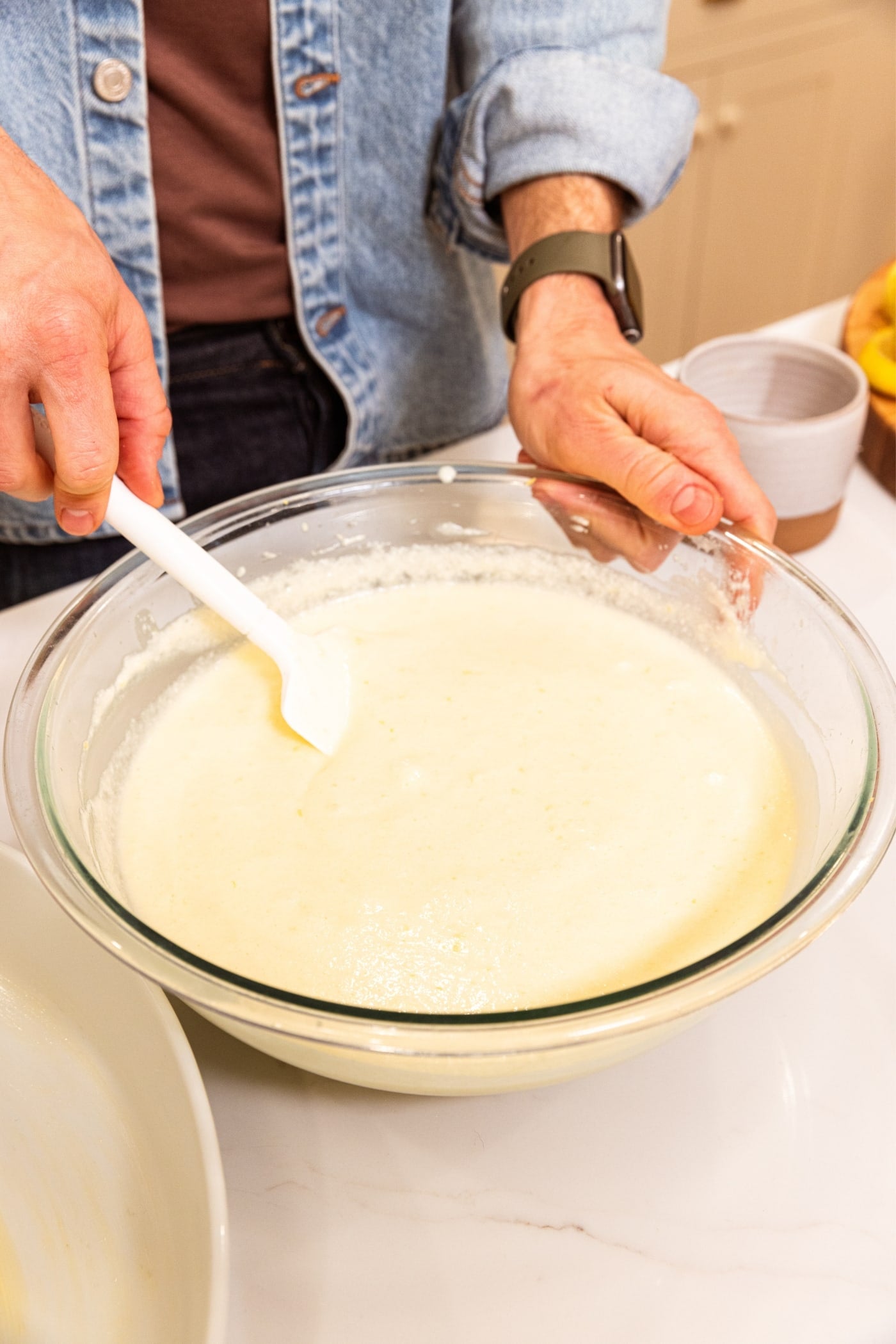 Hand holding white spatula gently folding together beaten egg whites with liquid batter until no lumps remain in large glass bowl.