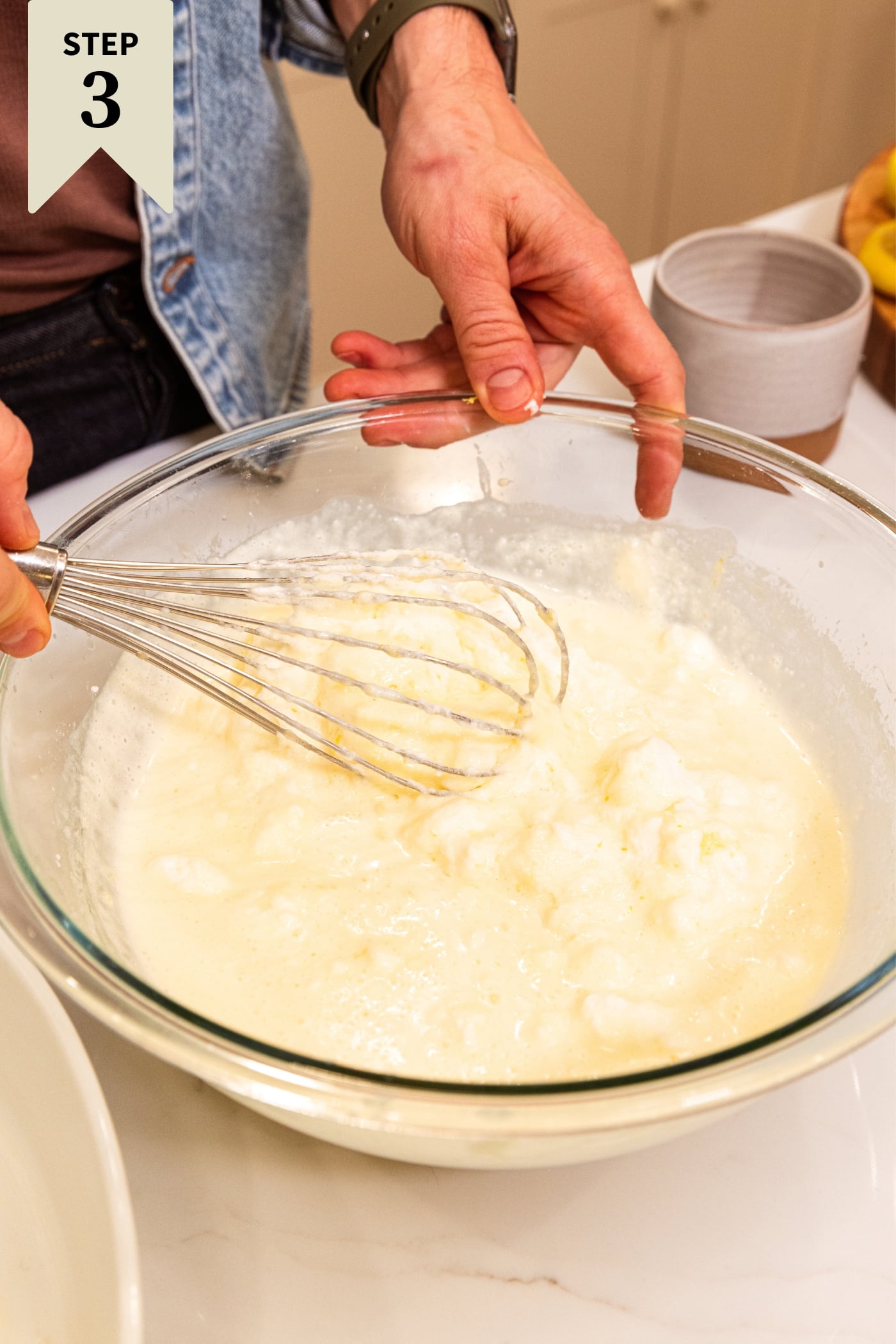 Hand using whisk to mix together beaten egg whites with a milky batter mixture in a large glass bowl on white surface.