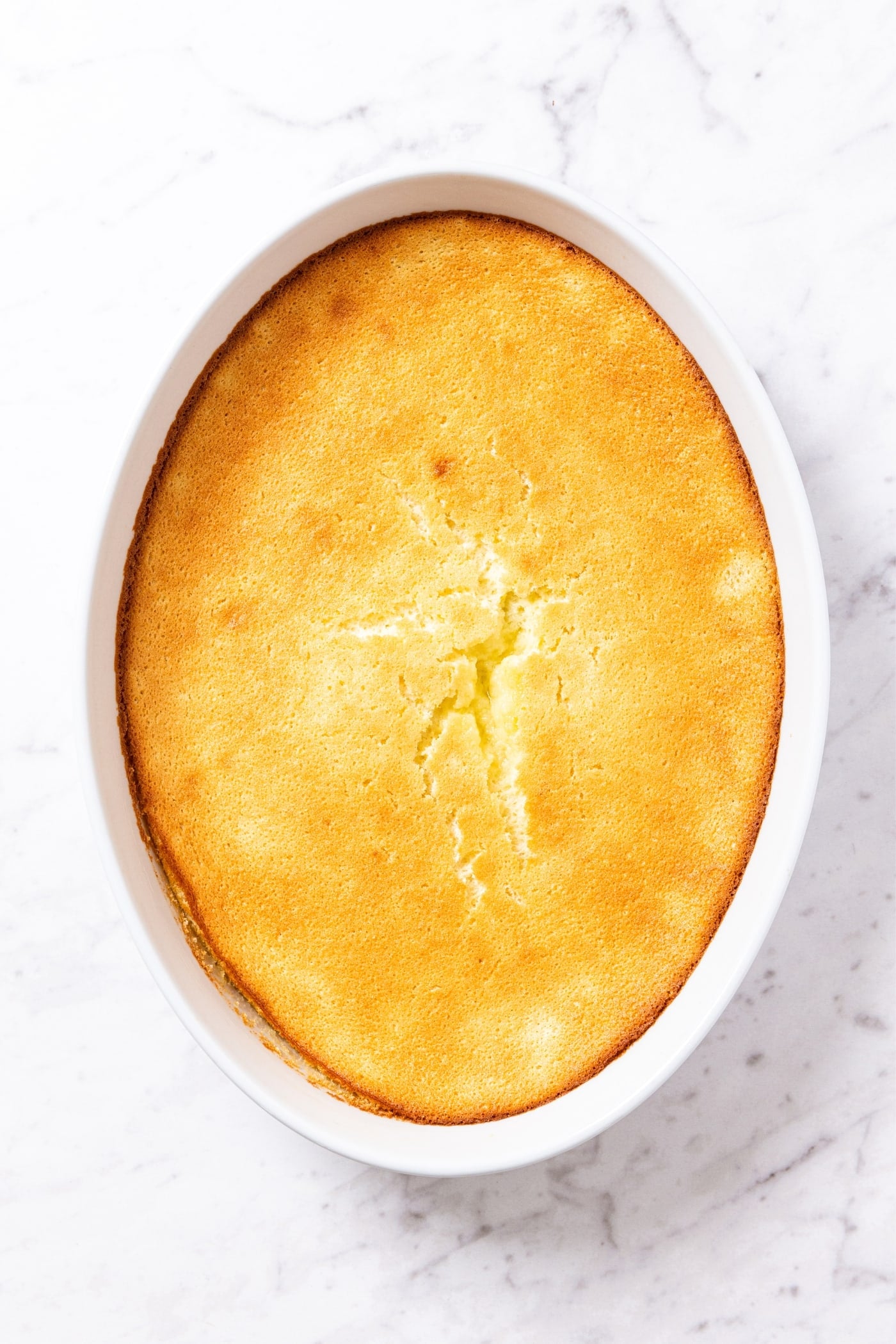 Large oval baking dish sitting on marble surface filled with baked lemon pudding cake with golden brown top crust and crack right down the middle.