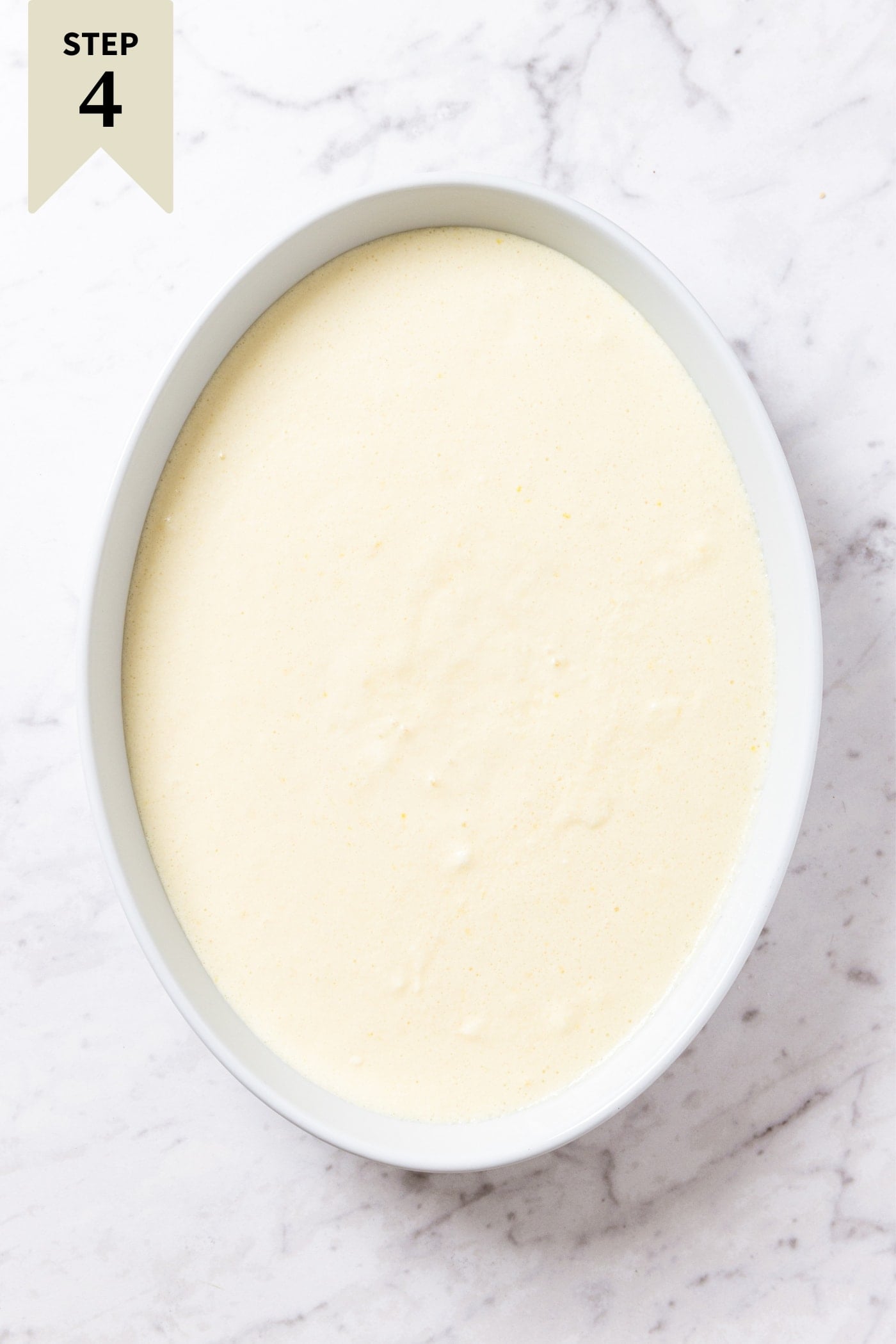 Large oval baking dish filled with pale yellow colored batter sitting on white marble surface right before going into the oven to bake.