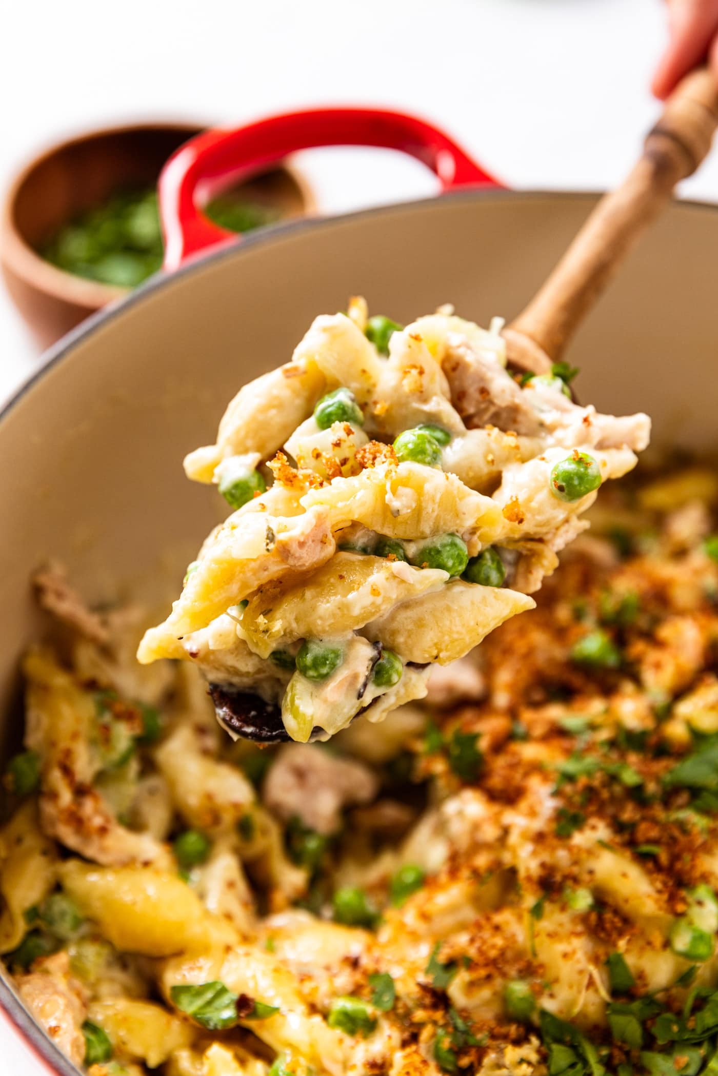 Wooden spoon holding serving of tuna noodle casserole with pot filled with rest of casserole below topped with browned bread crumbs.