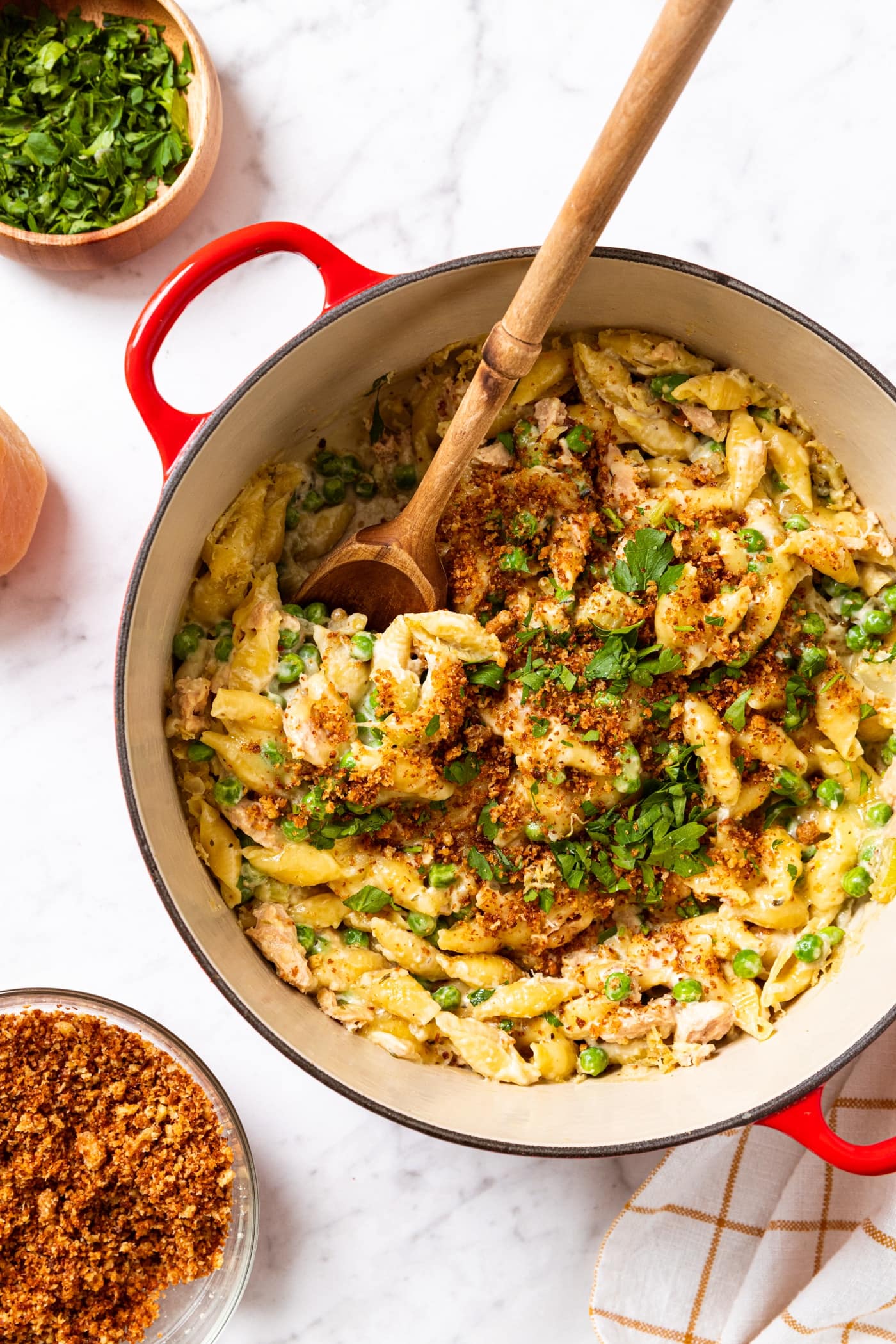 White marble surface with red Dutch oven filled with tuna noodle casserole with cheese melted on top with small bowl filled with bread crumbs.