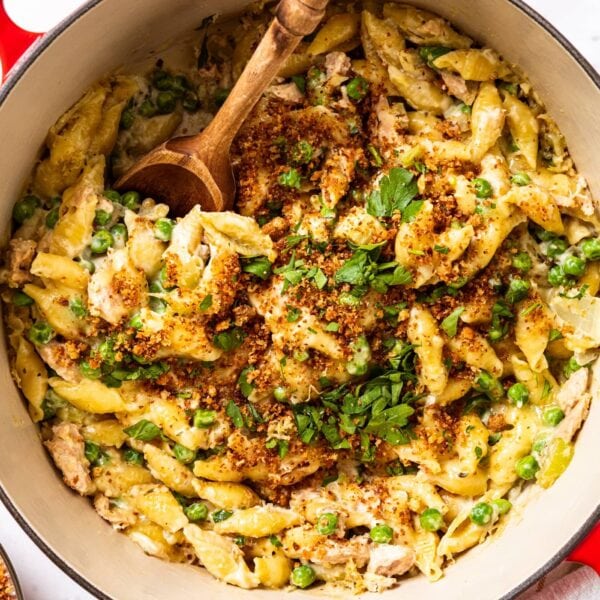 Large Dutch oven filled with tuna noodle casserole topped with melted cheese, chopped parsley, and browned bread crumbs with wooden spoon resting in bowl.